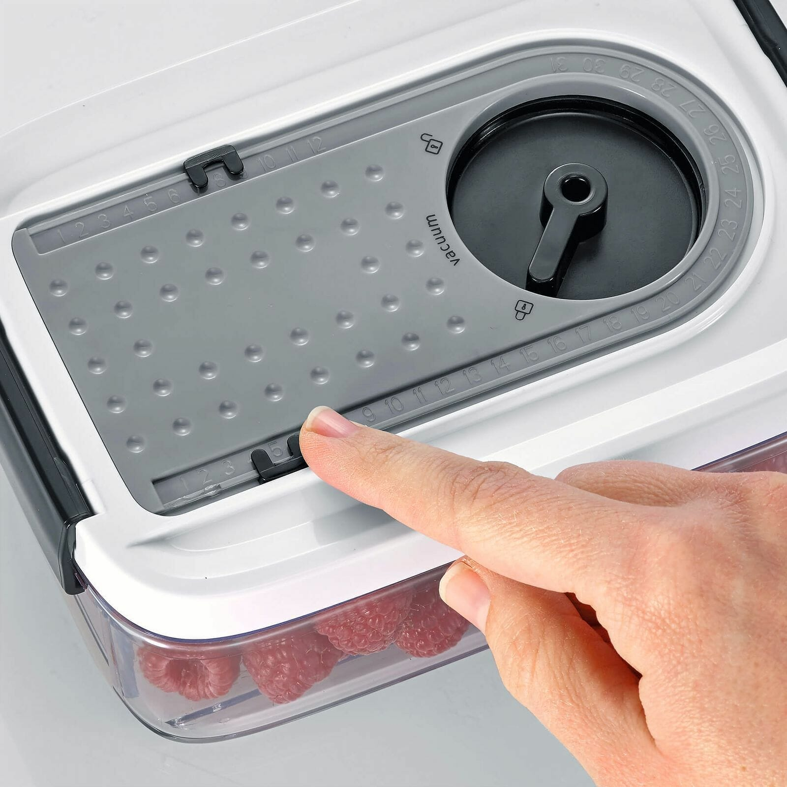 A hand presses a button on a food storage container lid, with raspberries visible inside.