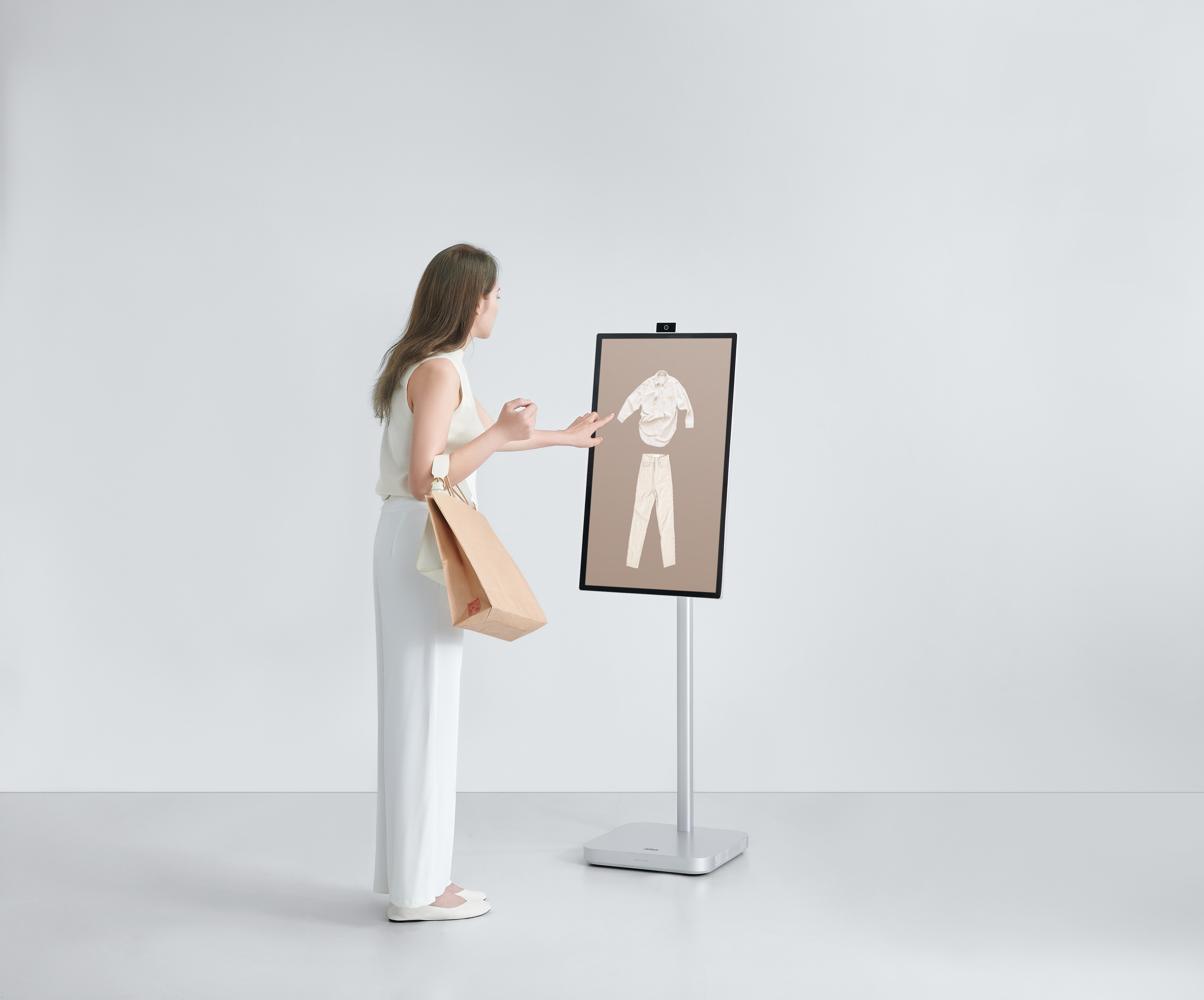 Kobieta patrzy na ekran pokazujący ubrania. Trzyma torbę. Stojak wyświetla ekran.