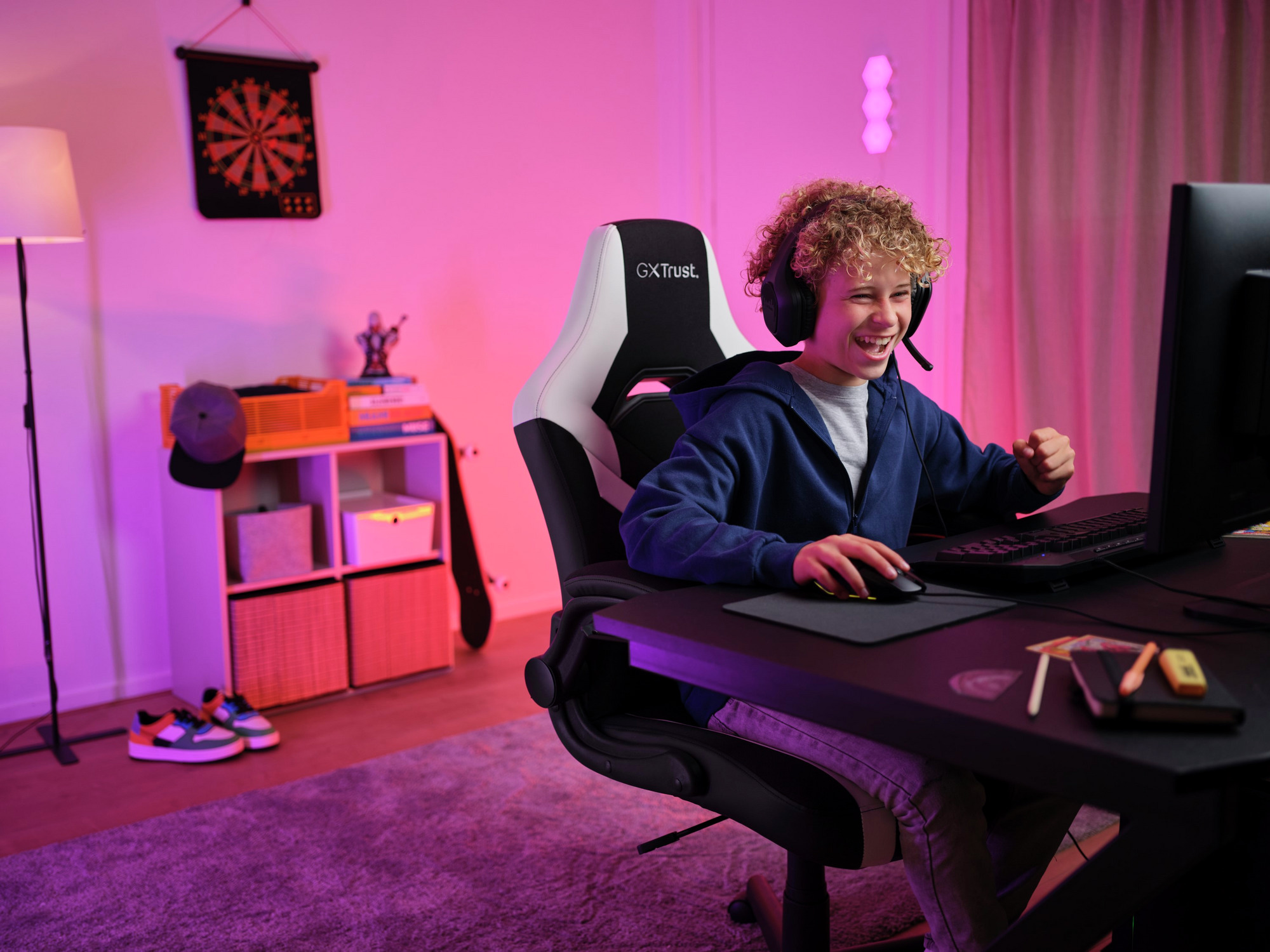 Junge spielt mit Headset, PC und Maus; Raum mit Dartscheibe und Lampe.