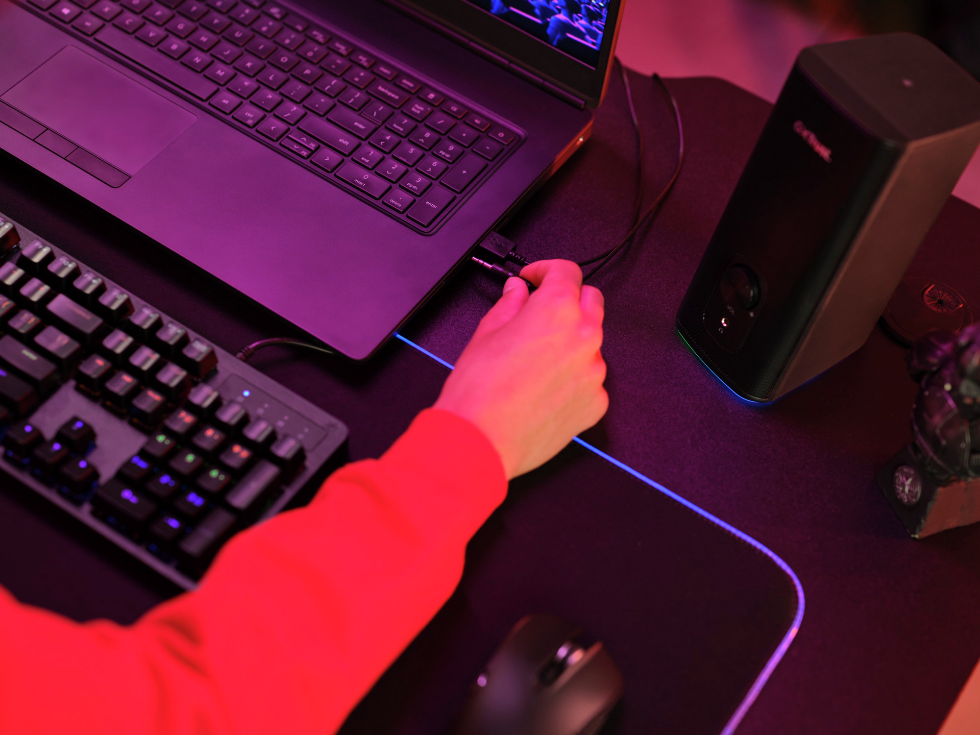 Osoba podłącza czarny kabel do laptopa i głośnika na biurku z myszką i klawiaturą.