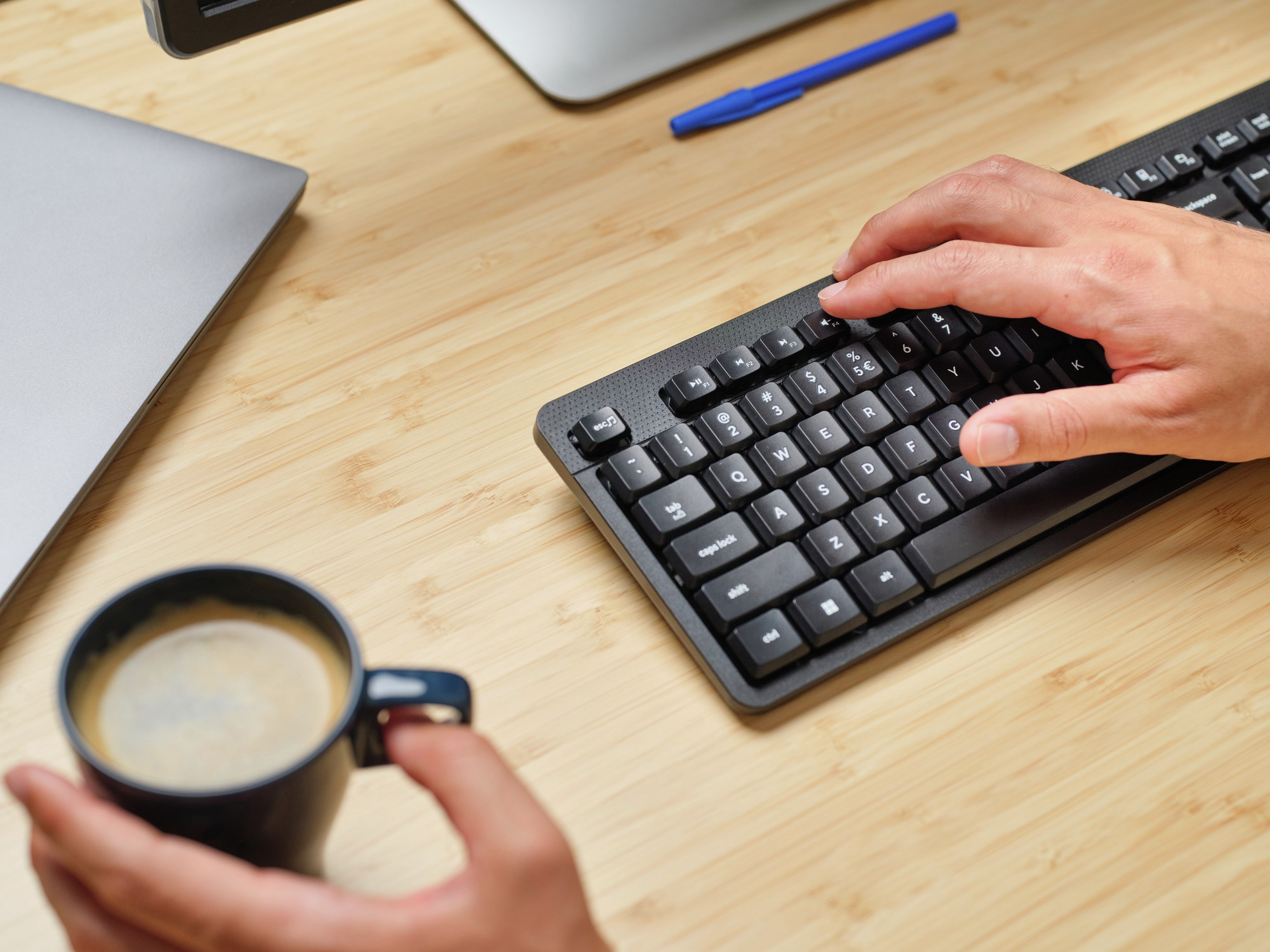 Ręce używające czarnej klawiatury obok filiżanki kawy i laptopa.