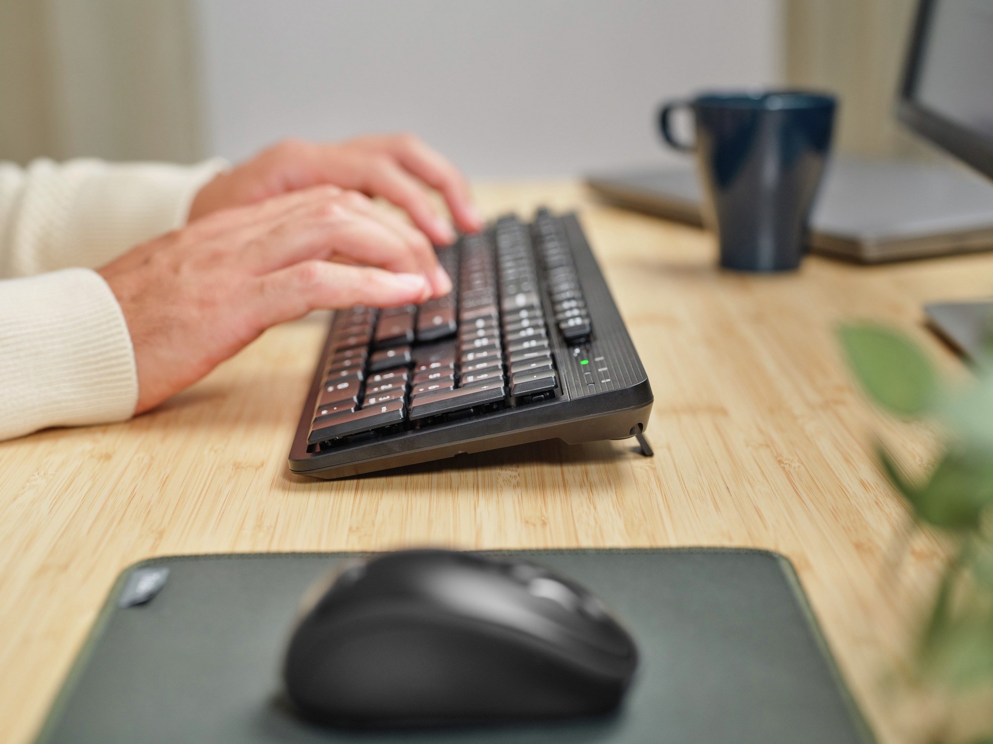 Dłonie piszące na czarnej klawiaturze, z myszą i kubkiem w pobliżu.