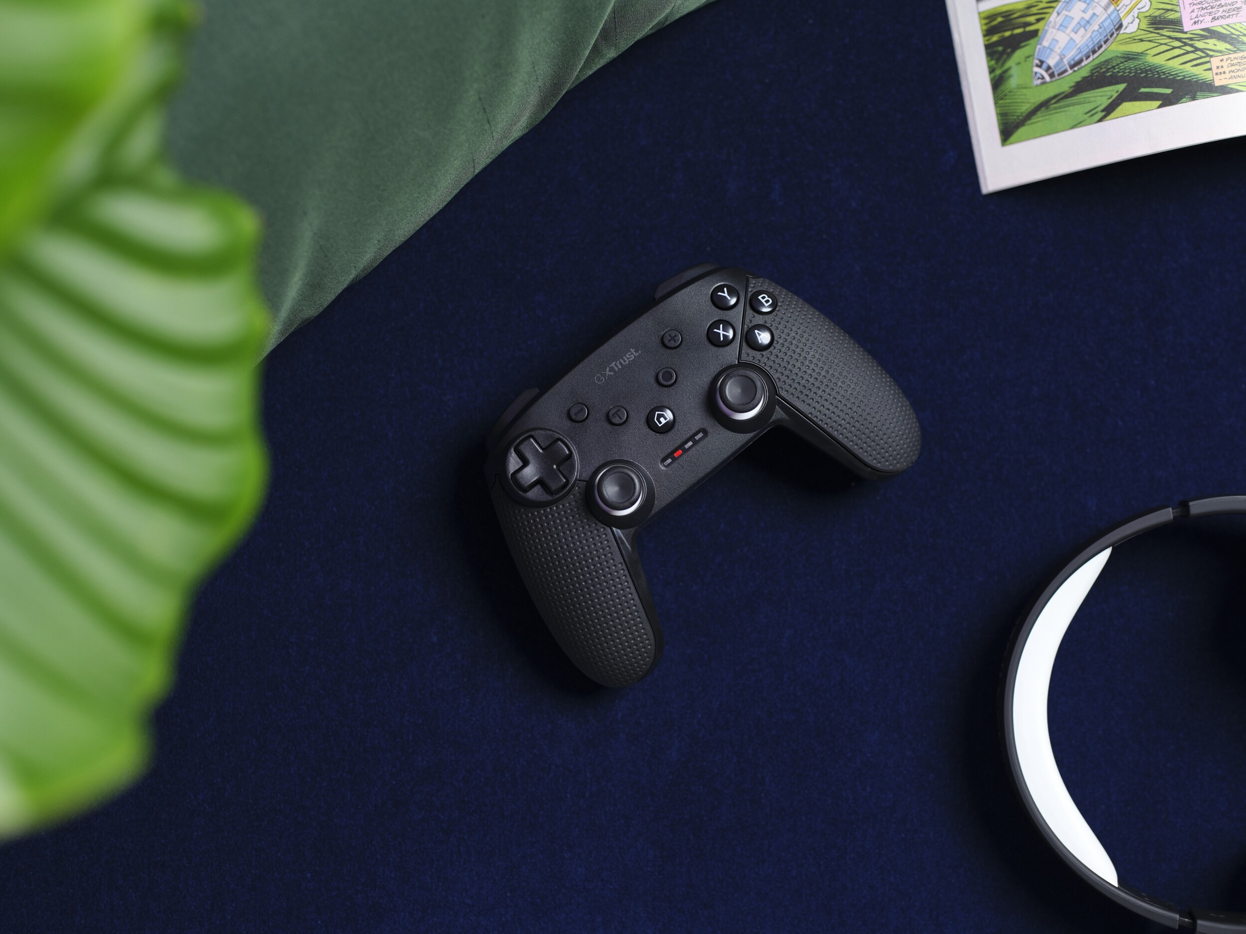Ein schwarzer Gaming-Controller auf dunkler Oberfläche, mit einer grünen Pflanze und einem weißen Kopfhörer an der Seite.