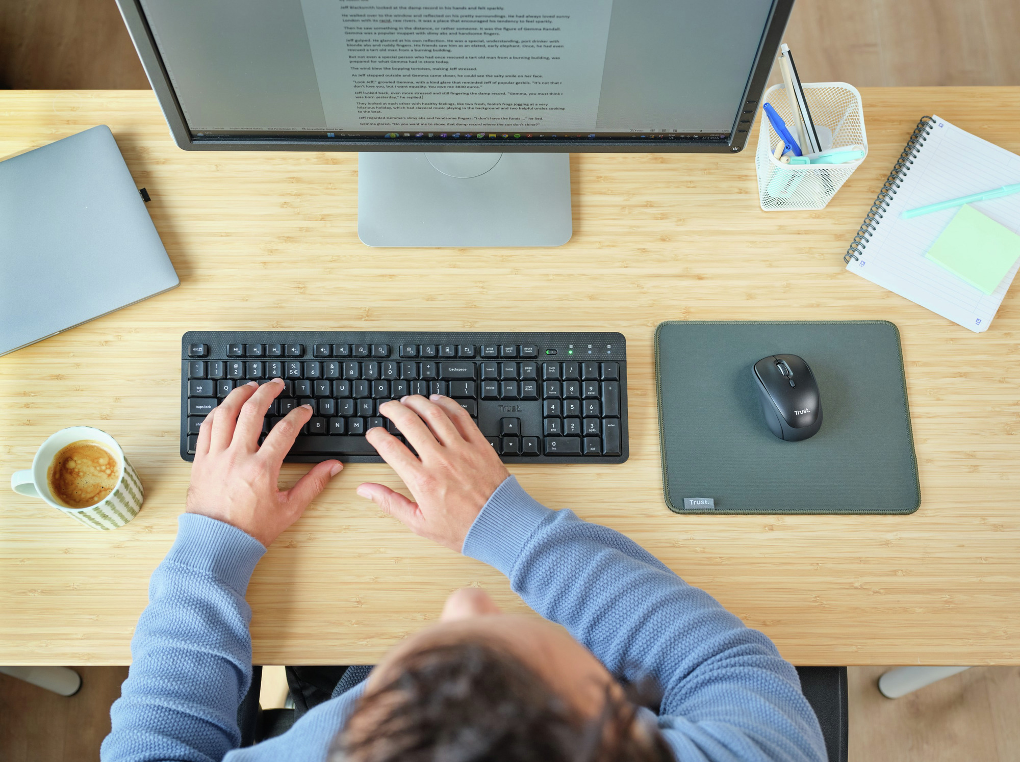 Widok z góry na dłonie piszące na klawiaturze, z myszą, monitorem i kawą na drewnianym biurku.