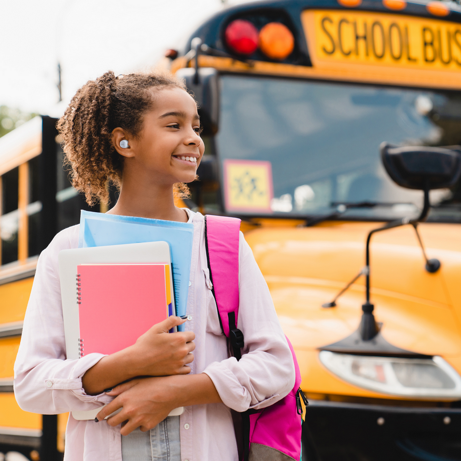 Dziewczyna uśmiechnięta przed żółtym autobusem szkolnym, trzymająca książki i plecak.