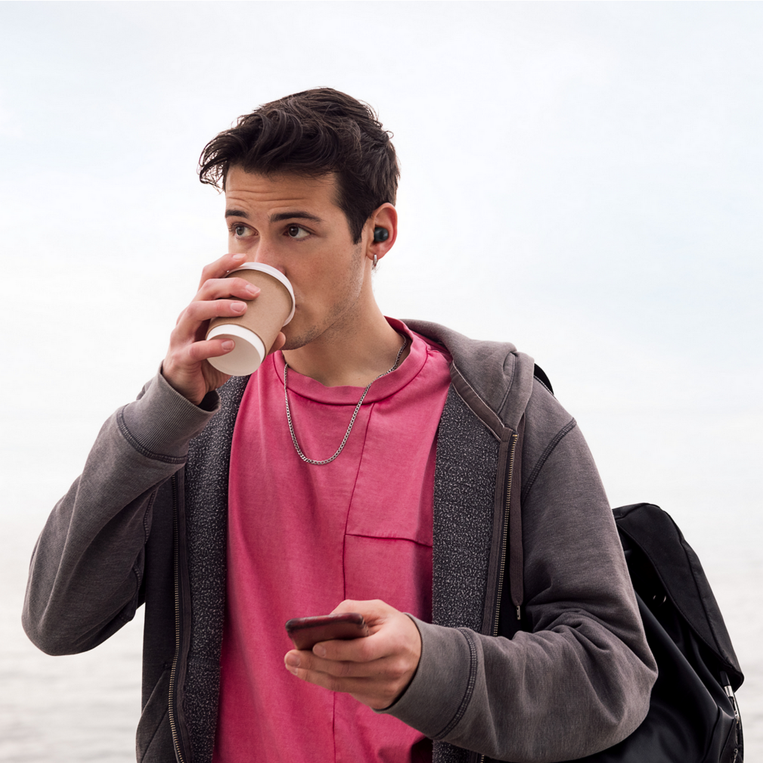 Mężczyzna pije z kubka kawy, ma na sobie słuchawki. Trzyma telefon i ma plecak.