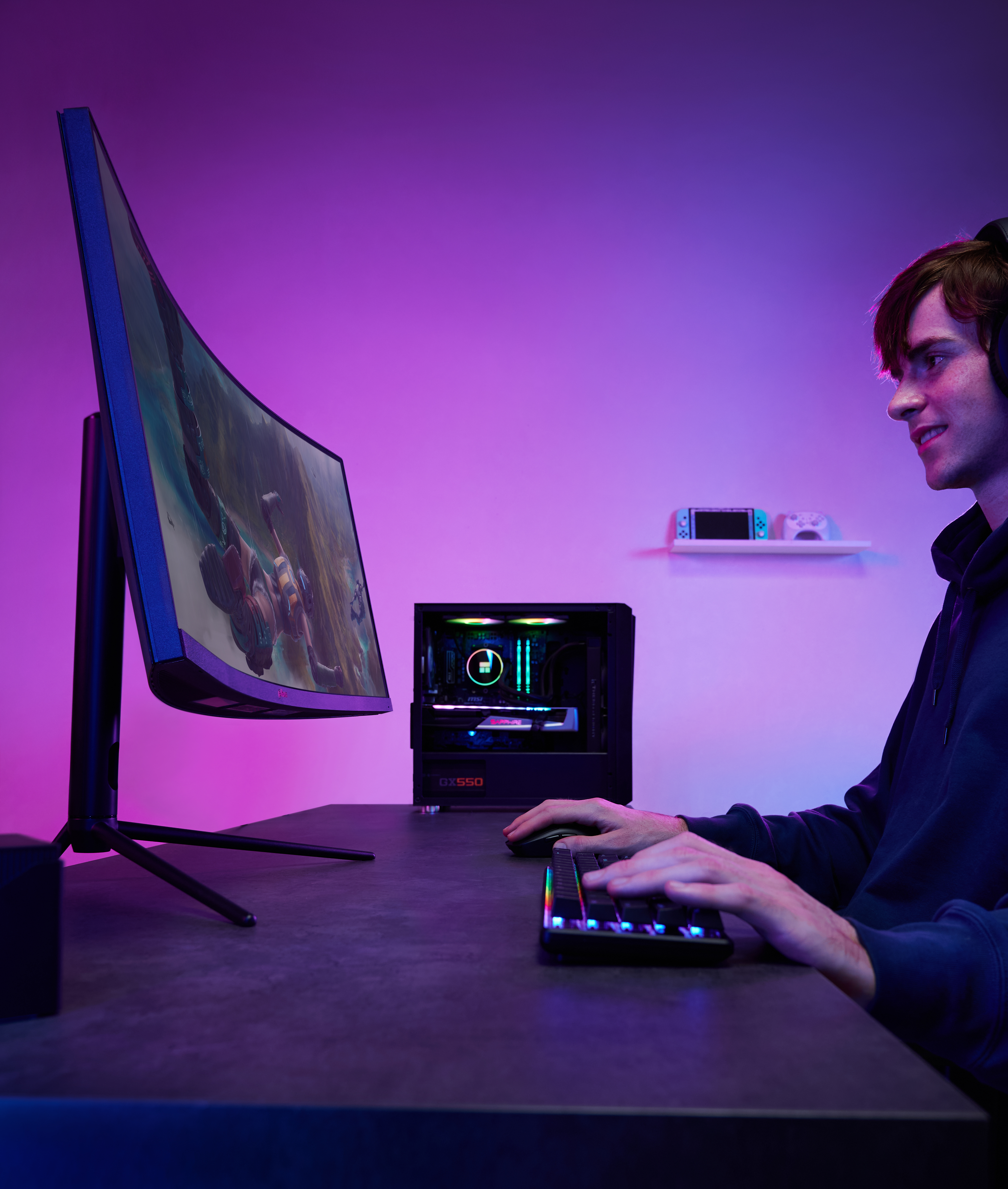 Gracz grający na komputerze. Zakrzywiony monitor, klawiatura z kolorowymi światłami, obudowa komputera. Fioletowo-różowe oświetlenie.