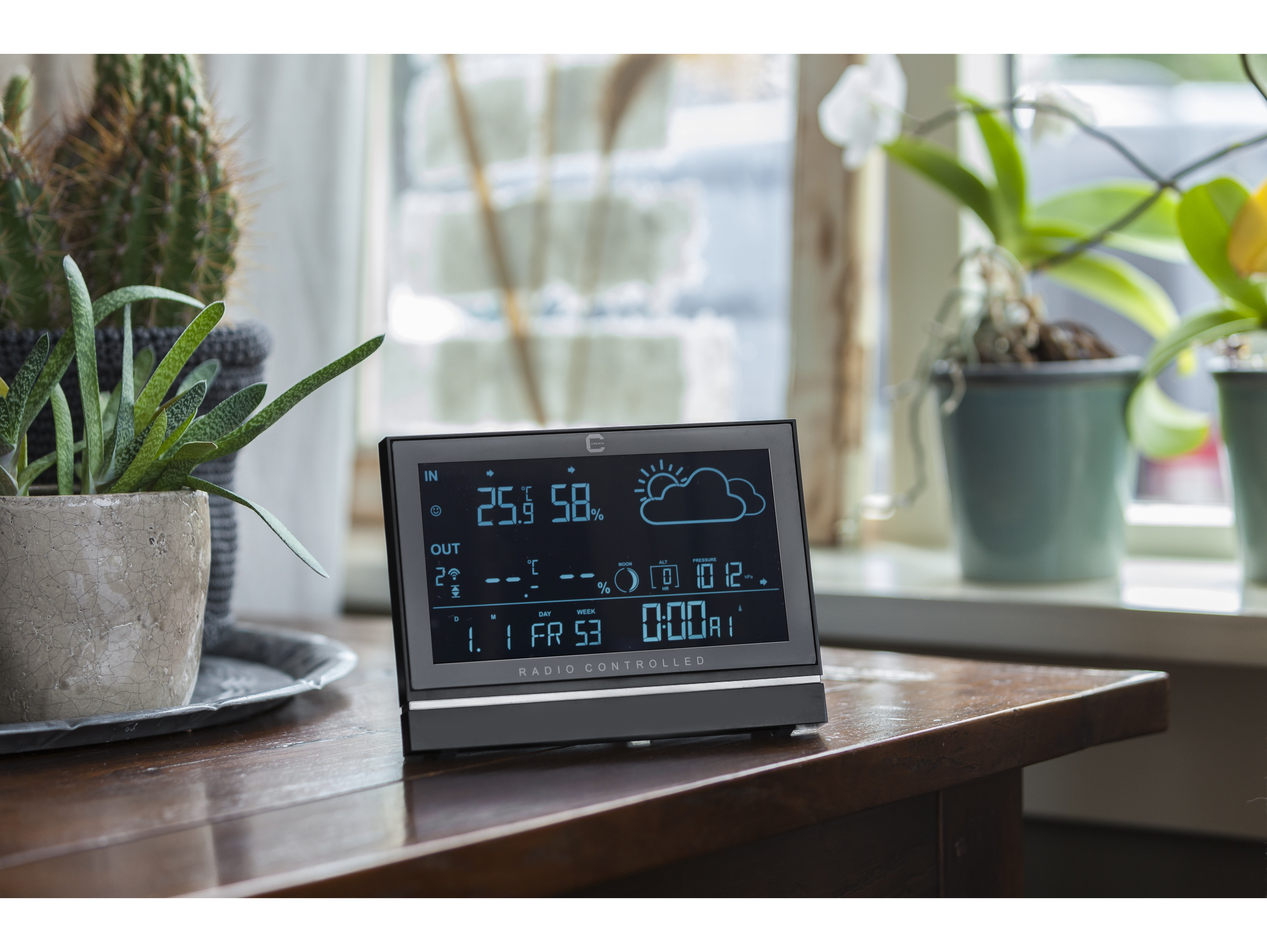 A digital weather station on a wooden table, with plants in the background.