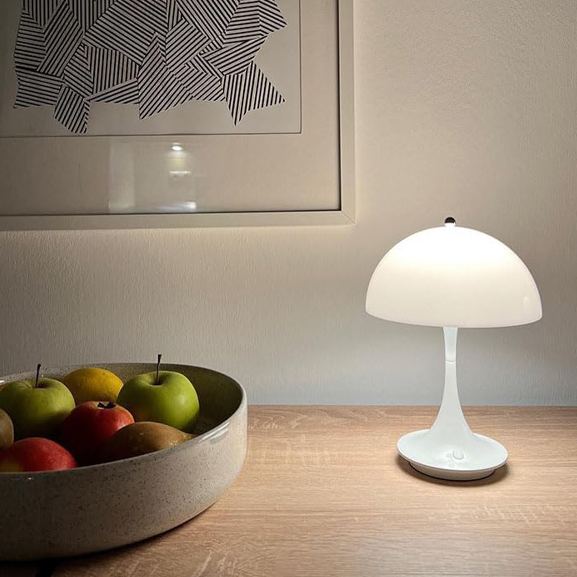 Weiße Tischlampe mit einer Obstschale auf einem Holztisch. Weißer Wandhintergrund.