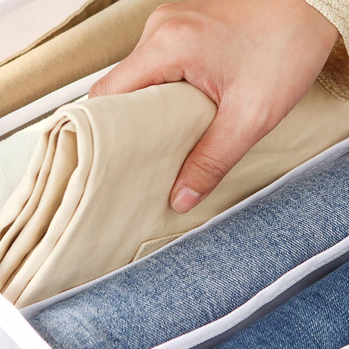 Eine Hand, die gefaltete beige Hose in einer Schublade mit Jeans und einem weißen Teiler hält.
