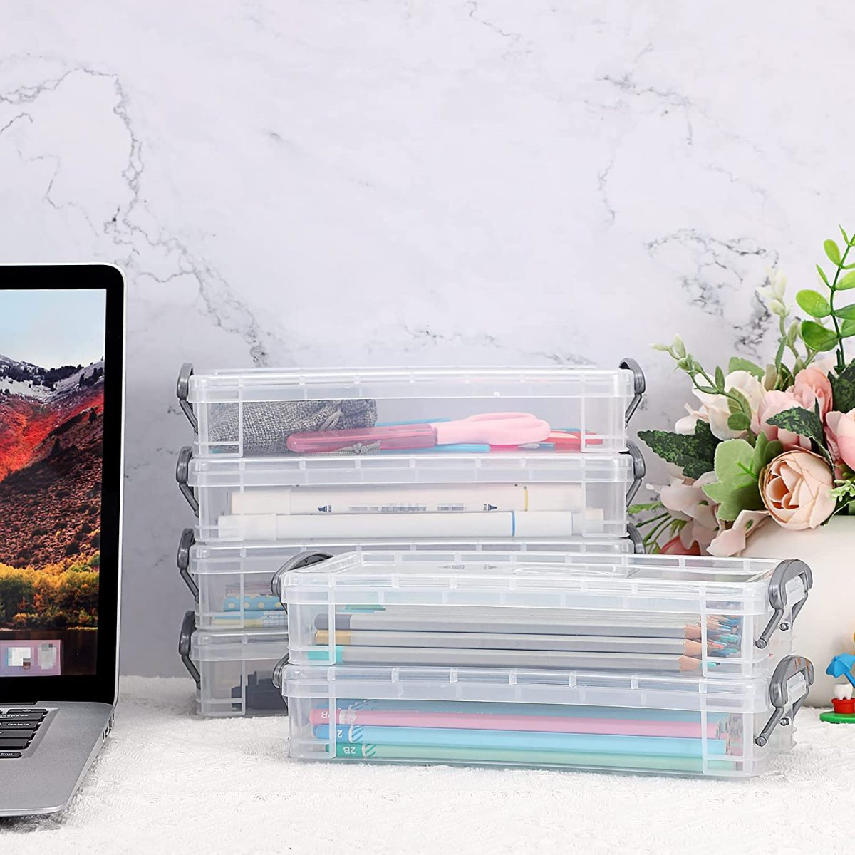 Mehrere transparente Stifteetuis gestapelt. Ein Laptop und Blumen im Hintergrund.
