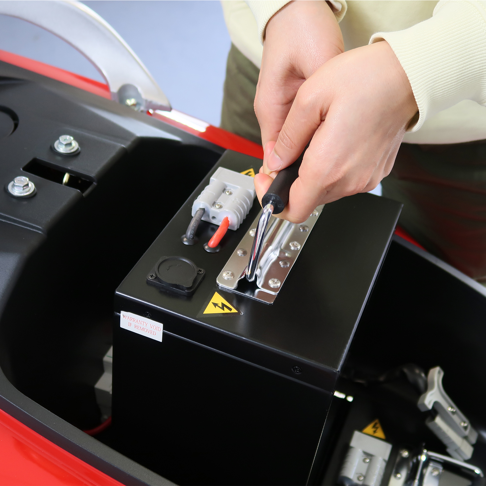 Hände einer Person, die ein Werkzeug an einer schwarzen Batterie in einem roten Fahrzeugfach verwenden.