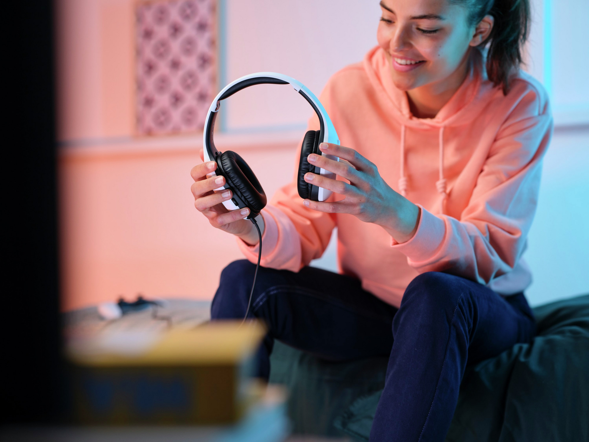 Kobieta trzyma biały zestaw słuchawkowy z czarnymi nausznikami, uśmiechnięta, w pokoju z niebieskim i pomarańczowym światłem.