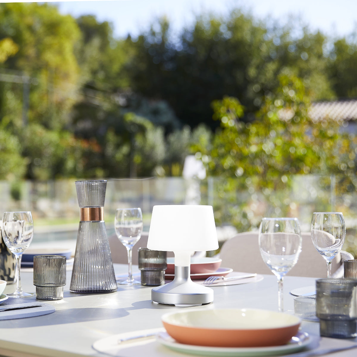 Esstisch im Freien mit Lampe, Gläsern und Essen, verschwommener Hintergrund mit Bäumen.