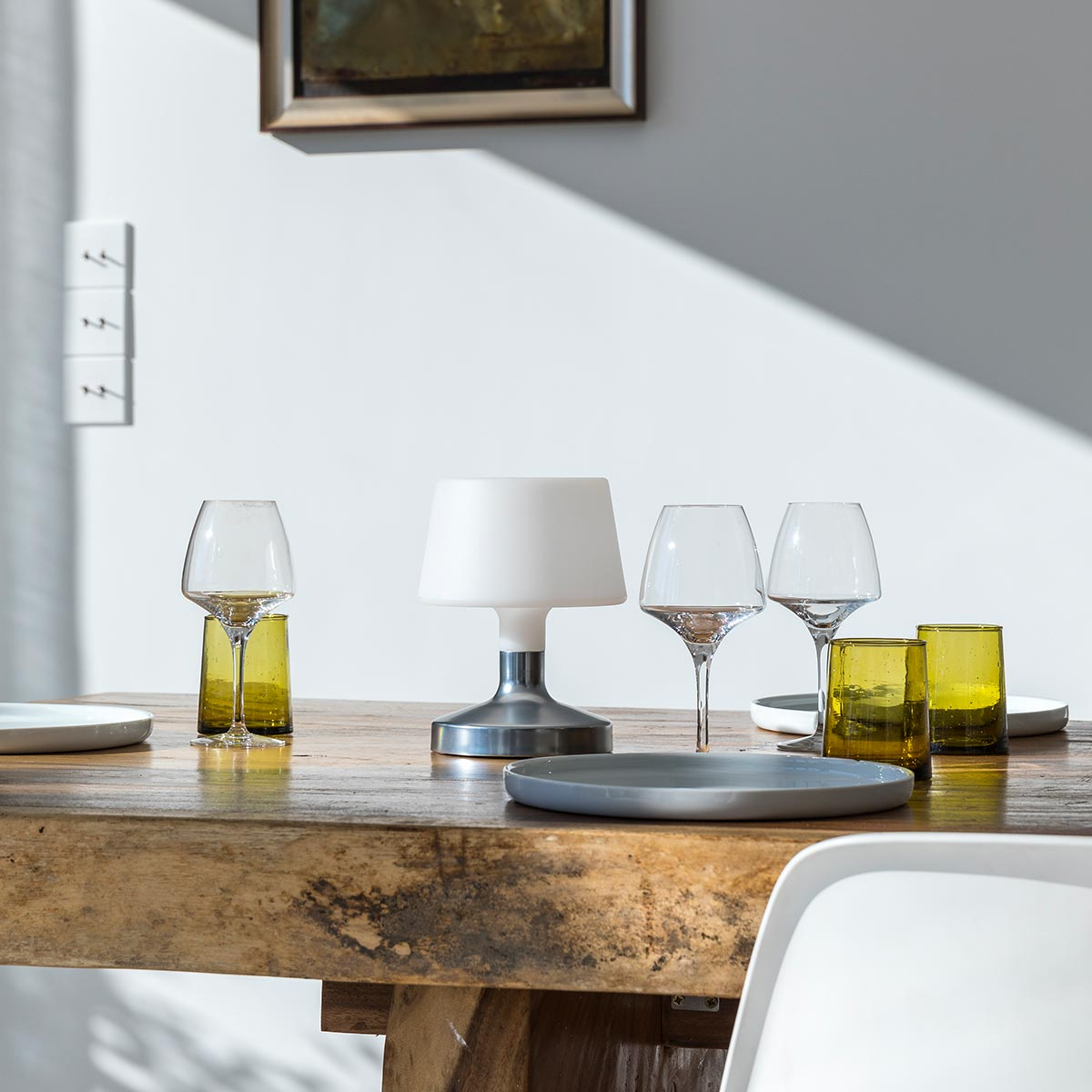 Tisch mit Weingläsern, Lampe und Tellern. Holztisch, weiße Wand im Hintergrund.