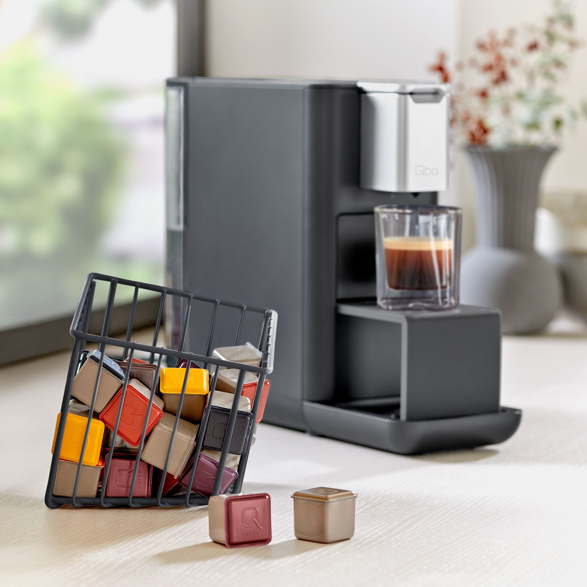 Eine dunkelgraue Kaffeemaschine mit einem Glas Kaffee und einem Korb mit Kaffeekapseln.