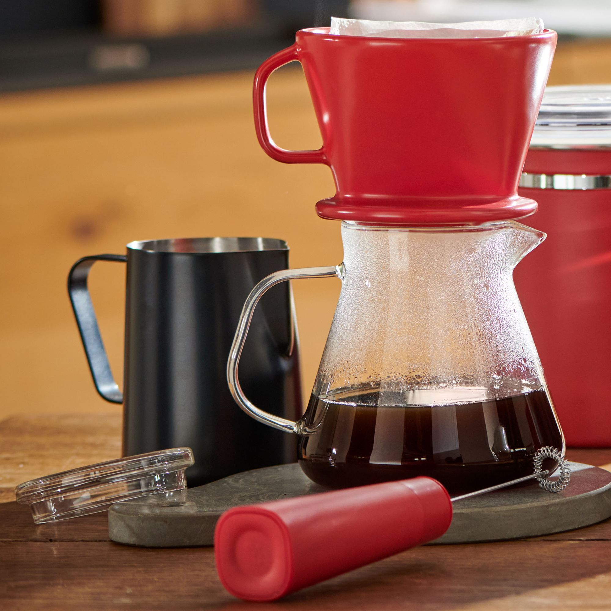 Eine rote Kaffeemaschine mit einem roten Pour-Over, einer schwarzen Kaffeekanne und einem roten Milchaufschäumer auf einem Holztisch.