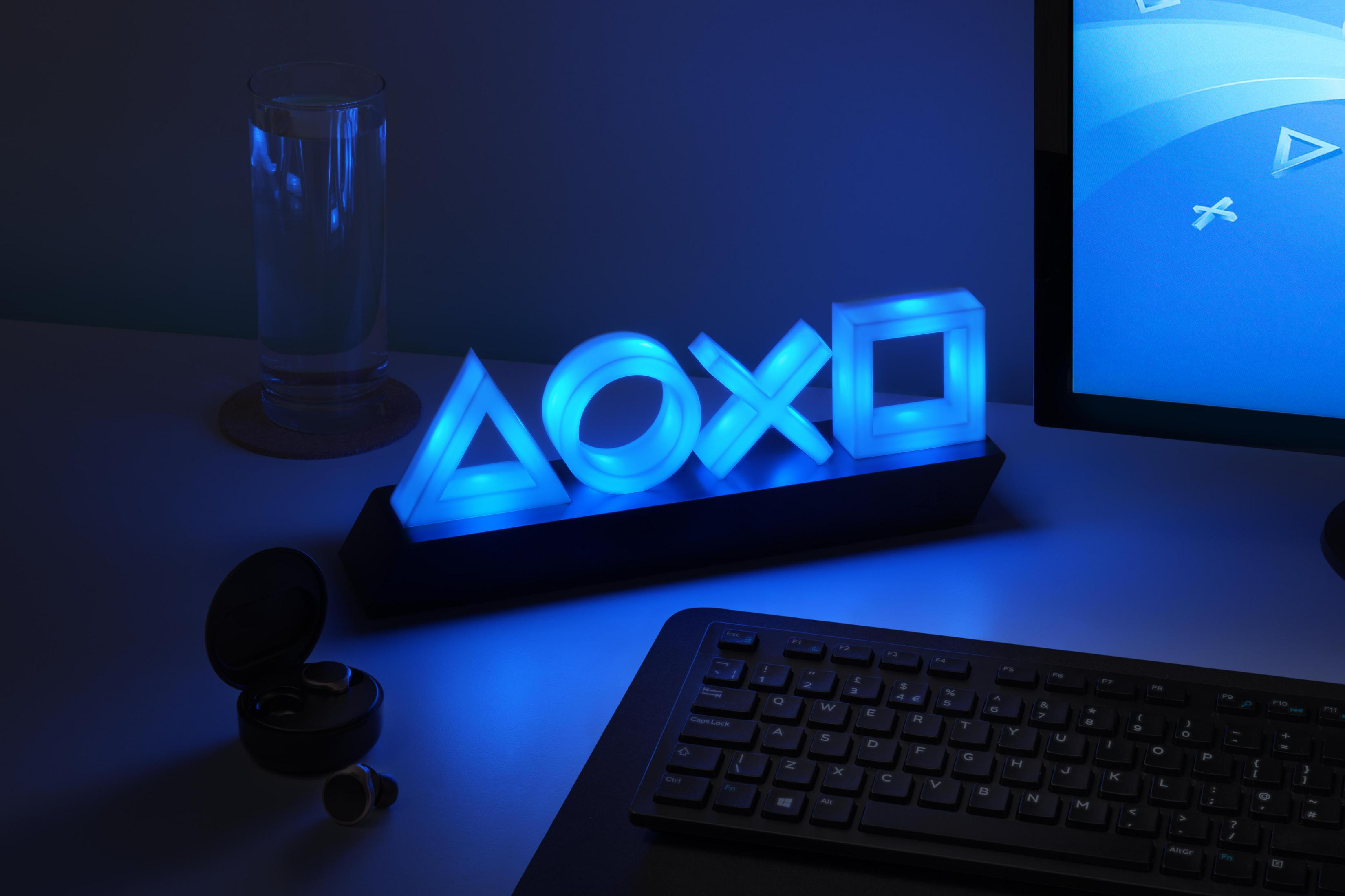 PlayStation-Symbol-Lampe mit blauen Lichtern auf einem Schreibtisch. Tastatur, Glas und Ohrhörer sind vorhanden.