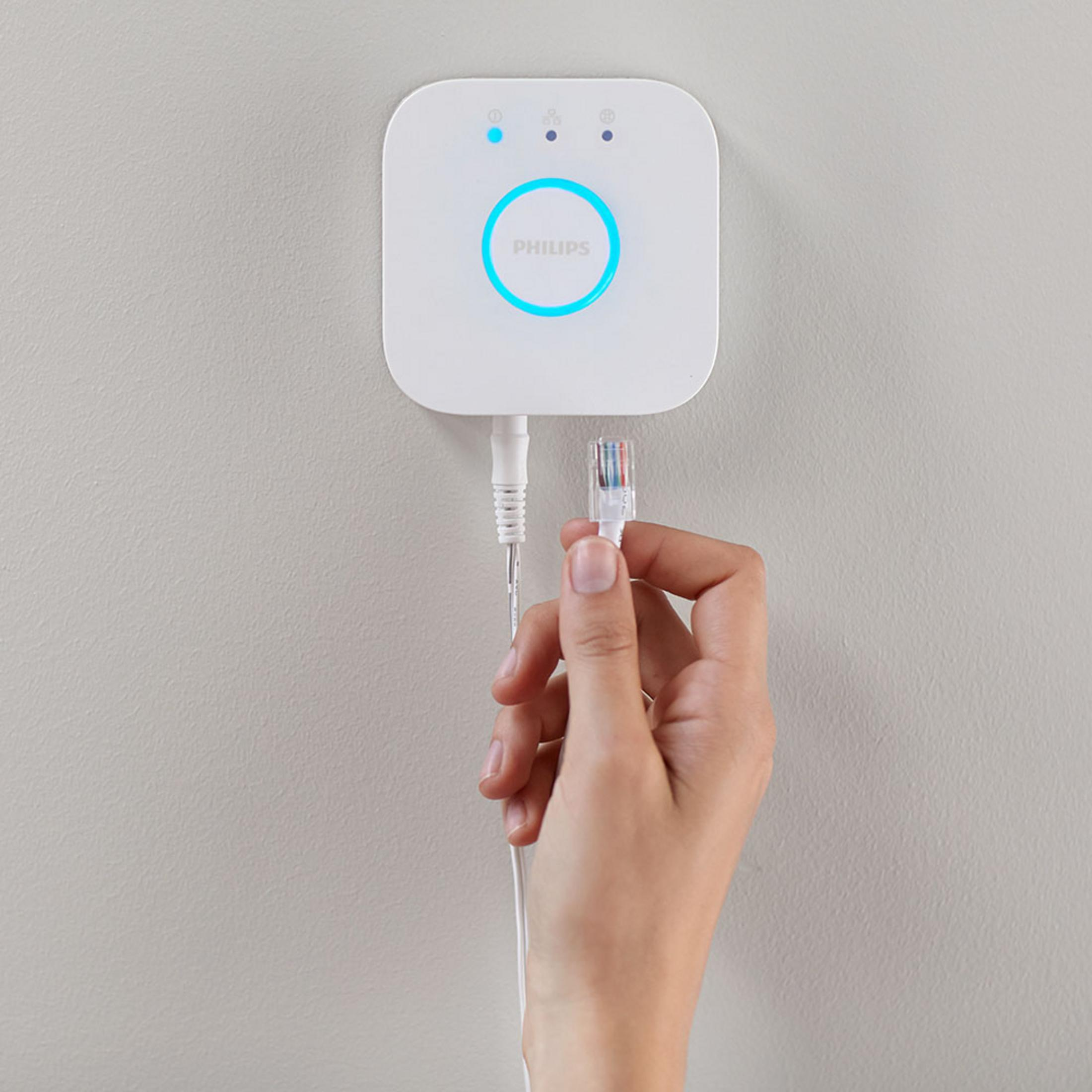 Weißer Philips Smart Home Hub mit einer Hand, die ein Kabel einsteckt, an einer weißen Wand.
