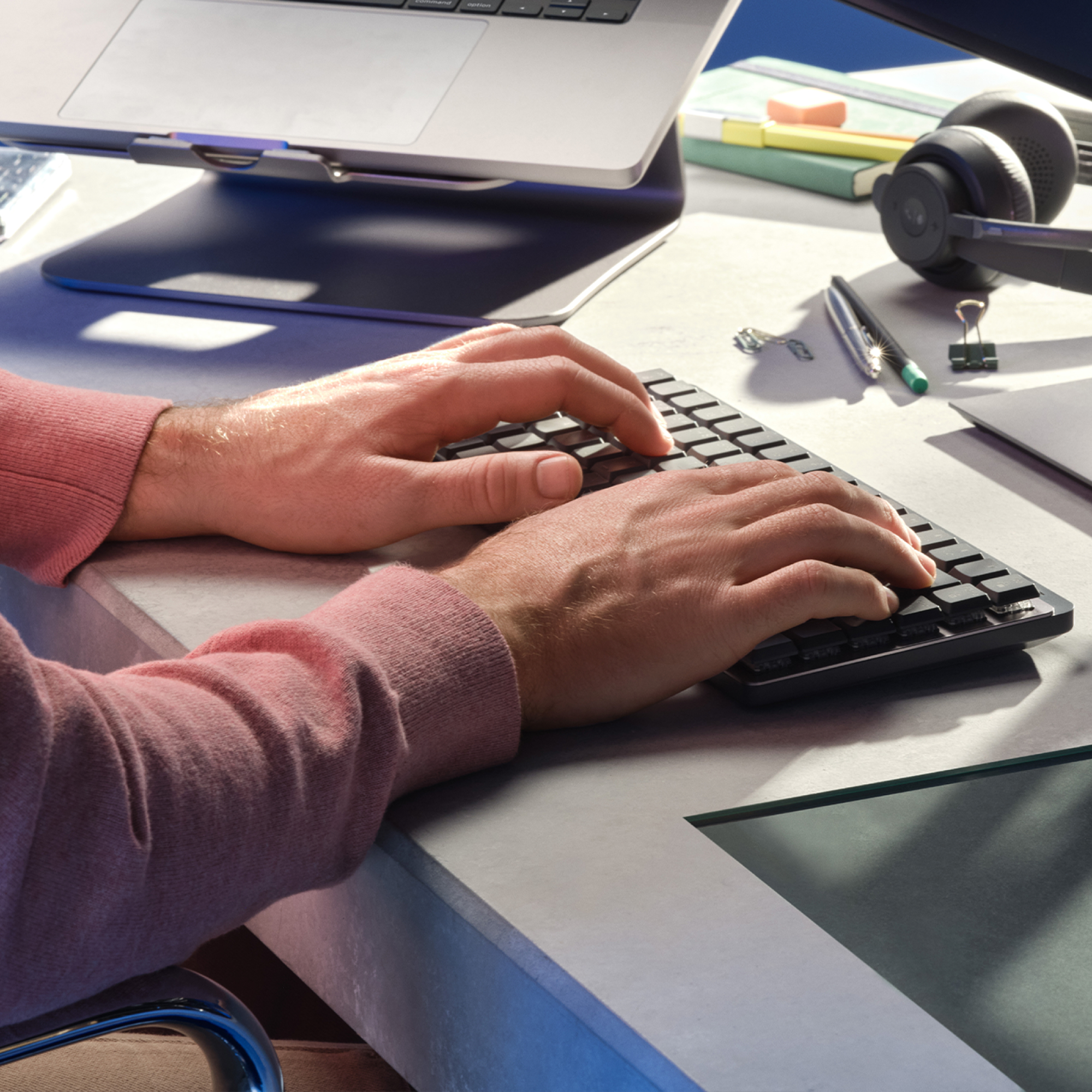 Dłonie piszące na czarnej klawiaturze na szarym biurku, z laptopem i słuchawkami w tle.