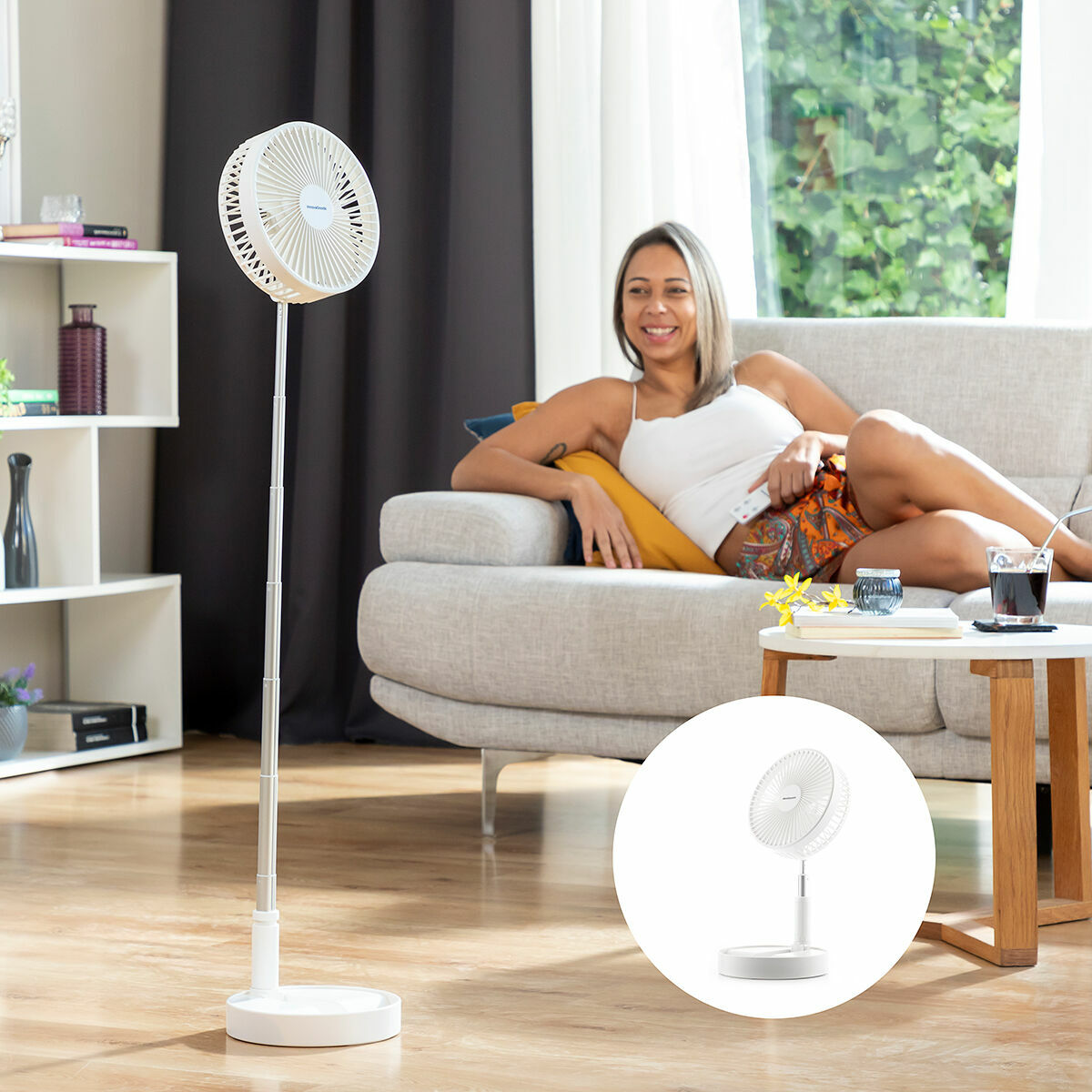 Mujer sentada en un sofá, ventilador blanco en el suelo. Mesa y ventana al fondo.