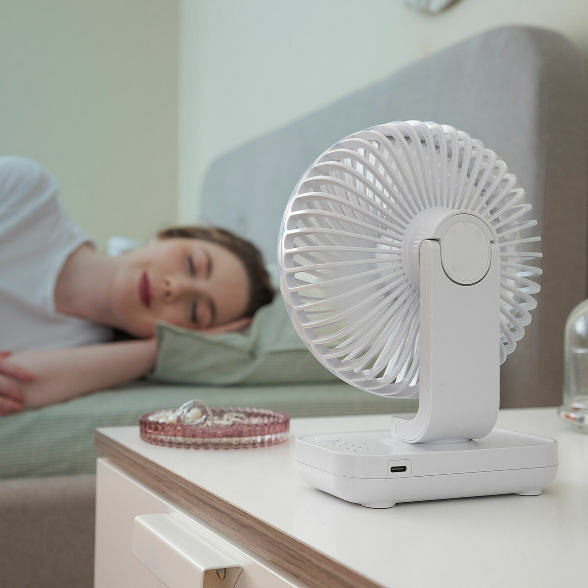 Ventilador blanco en mesita de noche, mujer durmiendo al fondo.
