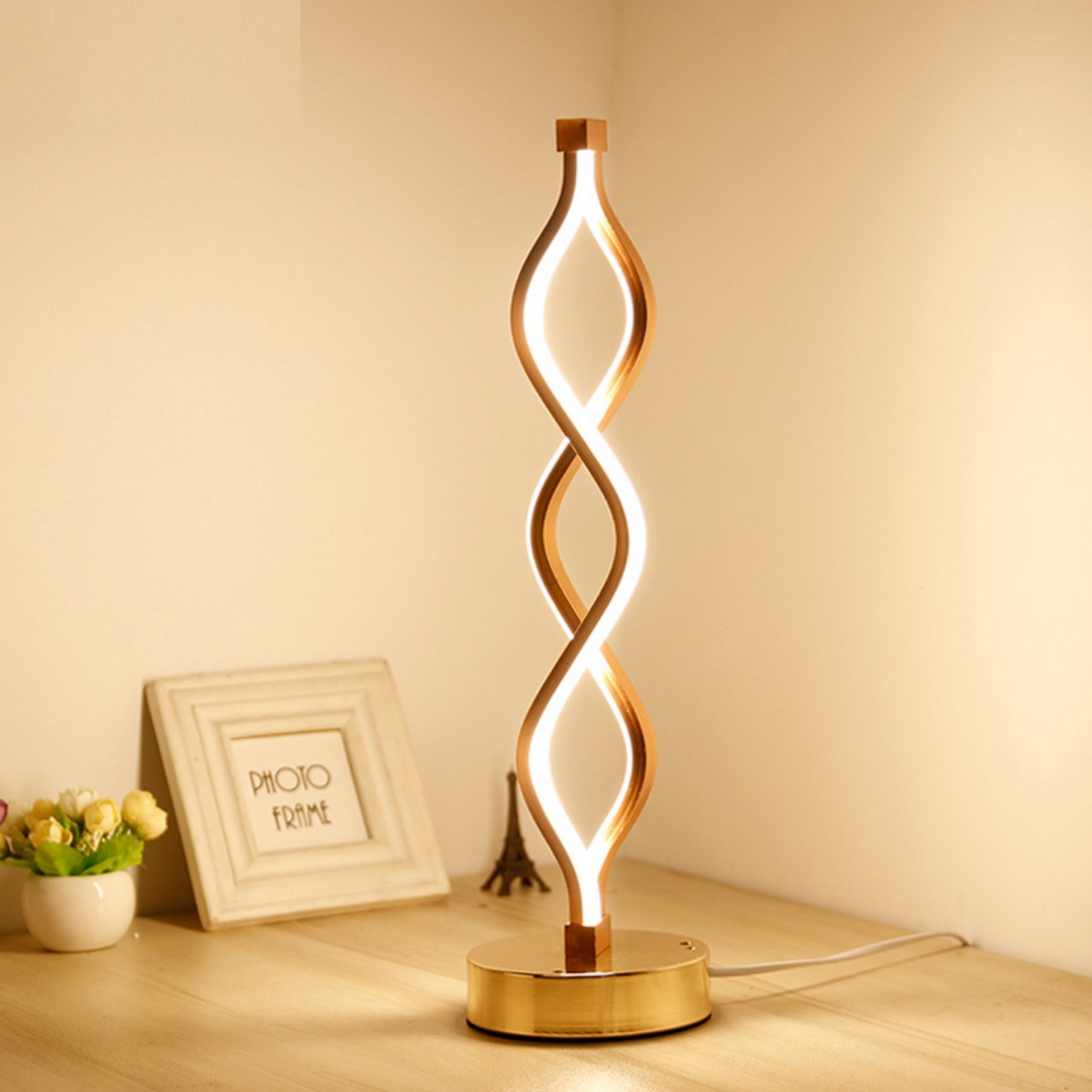 Moderne Lampe mit goldenem Sockel und gewundener Leuchte. Der Hintergrund ist eine beige Wand.