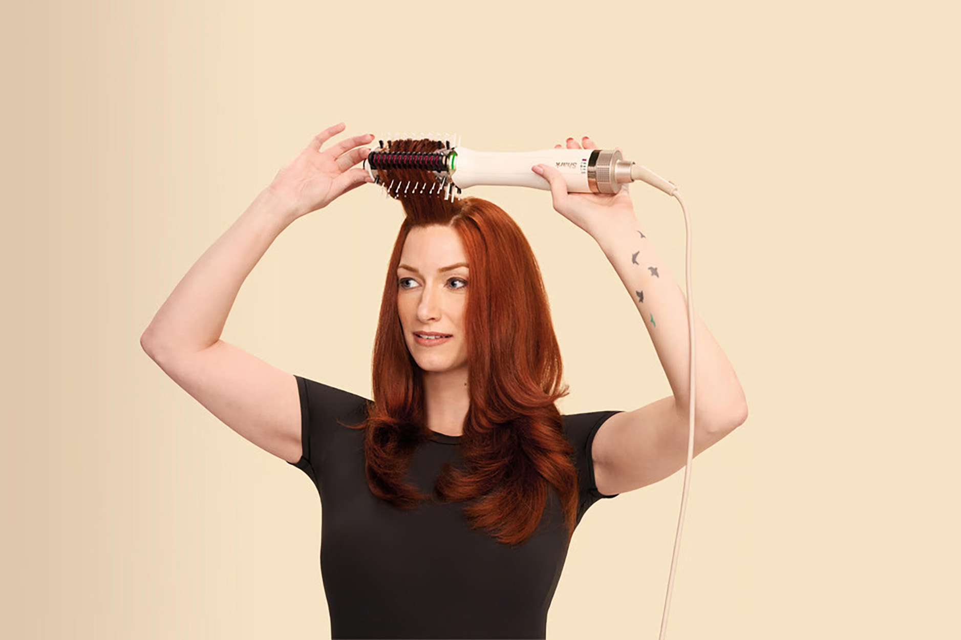 Kobieta używająca szczotki do włosów. Ma rude włosy, czarną koszulę, beżowe tło.