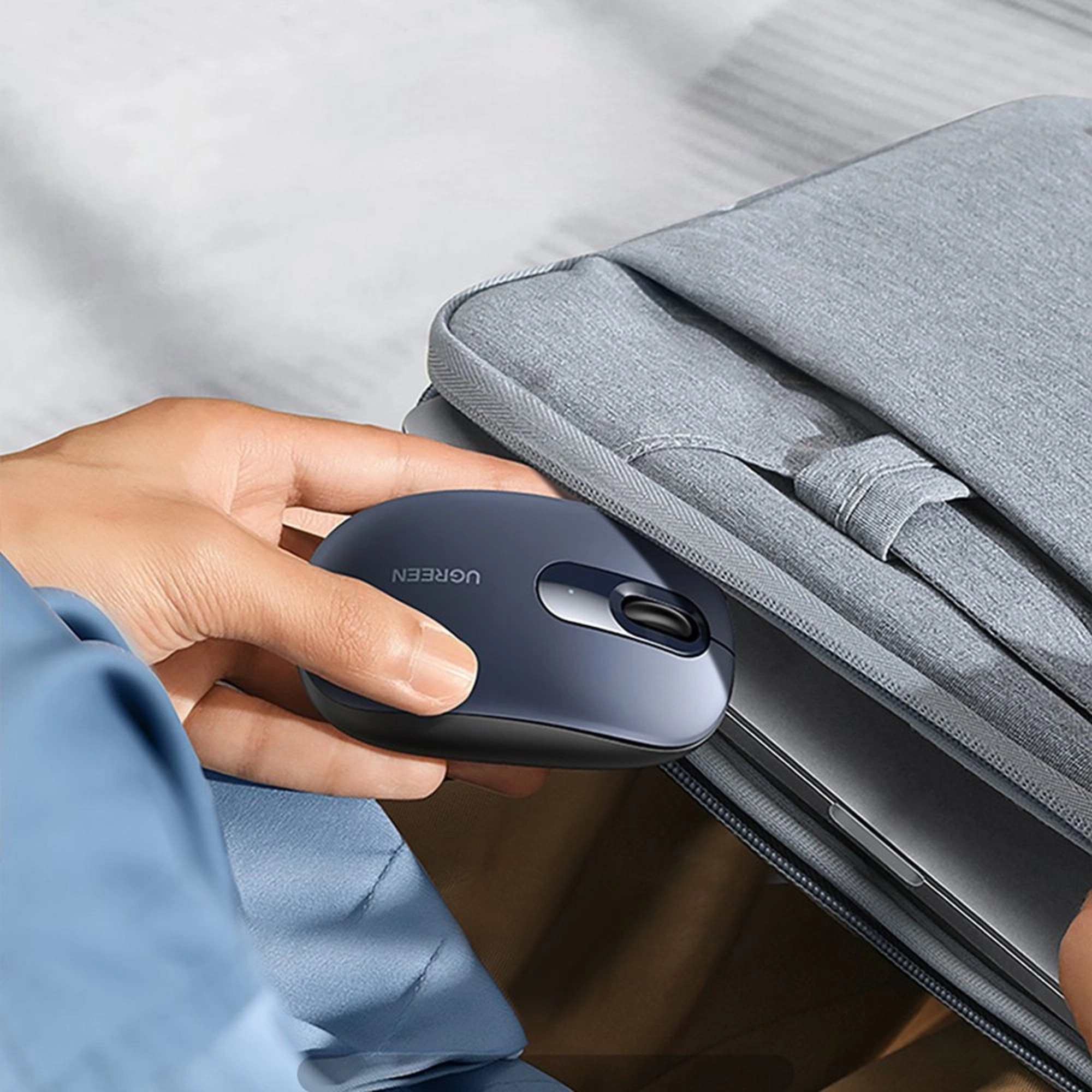 Die Hand einer Person, die eine dunkelblaue kabellose UGREEN-Maus hält, neben einer grauen Laptoptasche.