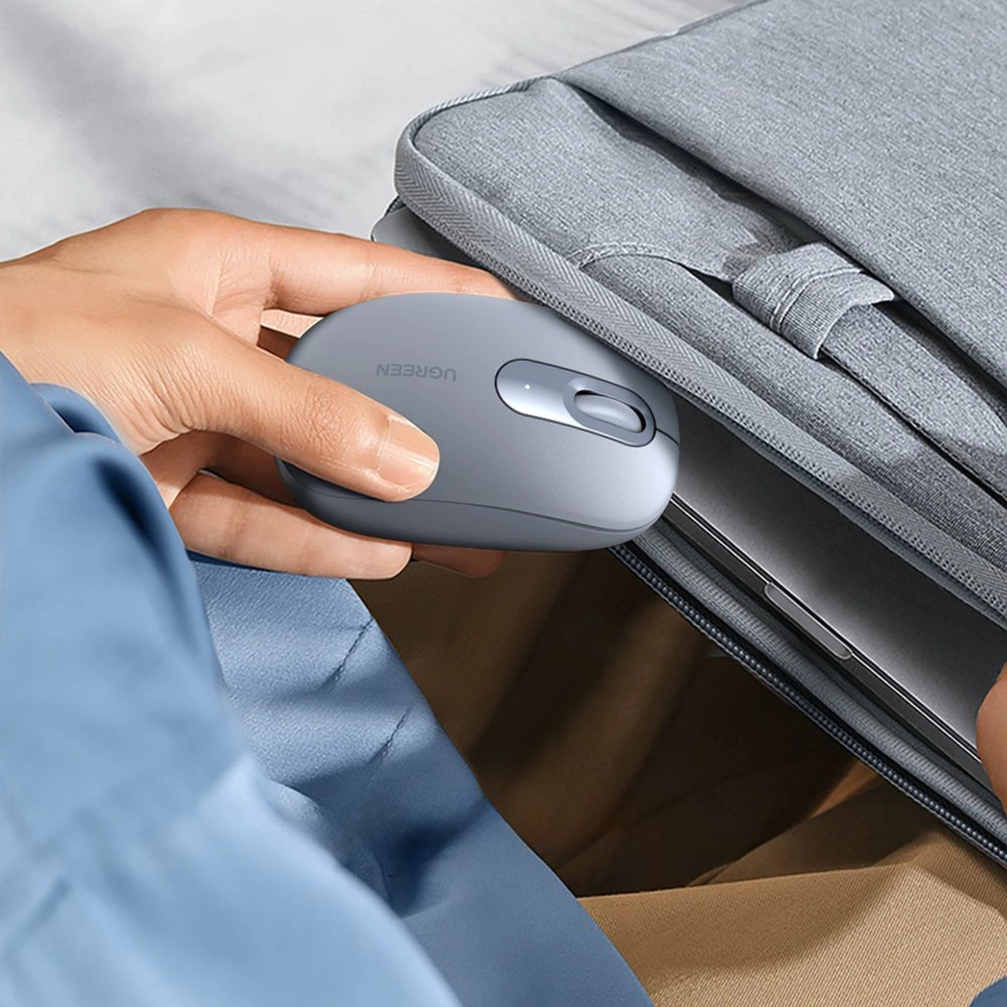 Hand hält graue Computermaus, mit grauem Laptop-Etui im Hintergrund.