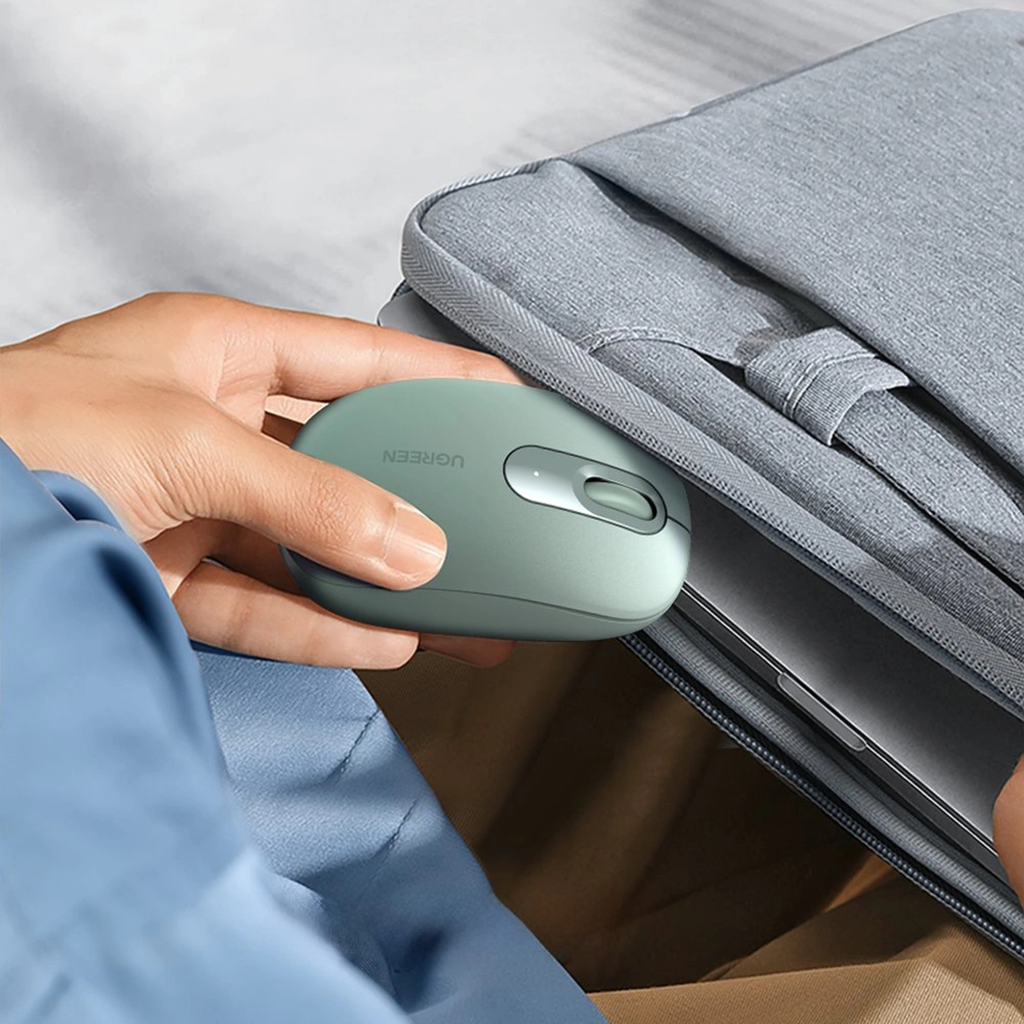 Eine hellgrüne UGREEN-Maus, die von einer Hand neben einer grauen Laptop-Hülle gehalten wird, auf neutralem Hintergrund.