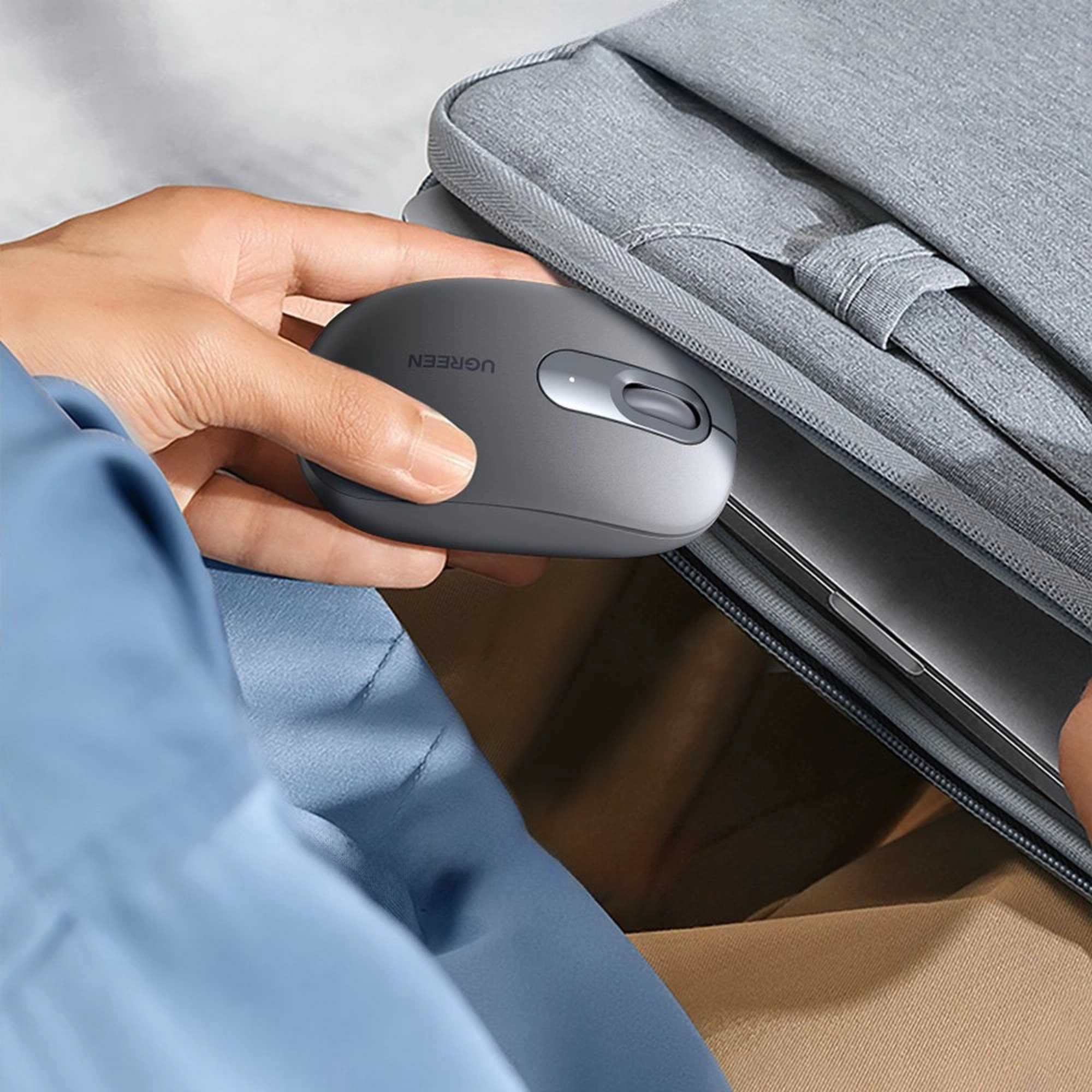 Eine Hand hält eine graue UGREEN-Maus in der Nähe eines grauen Laptop-Etuis. Der Hintergrund ist unscharf.
