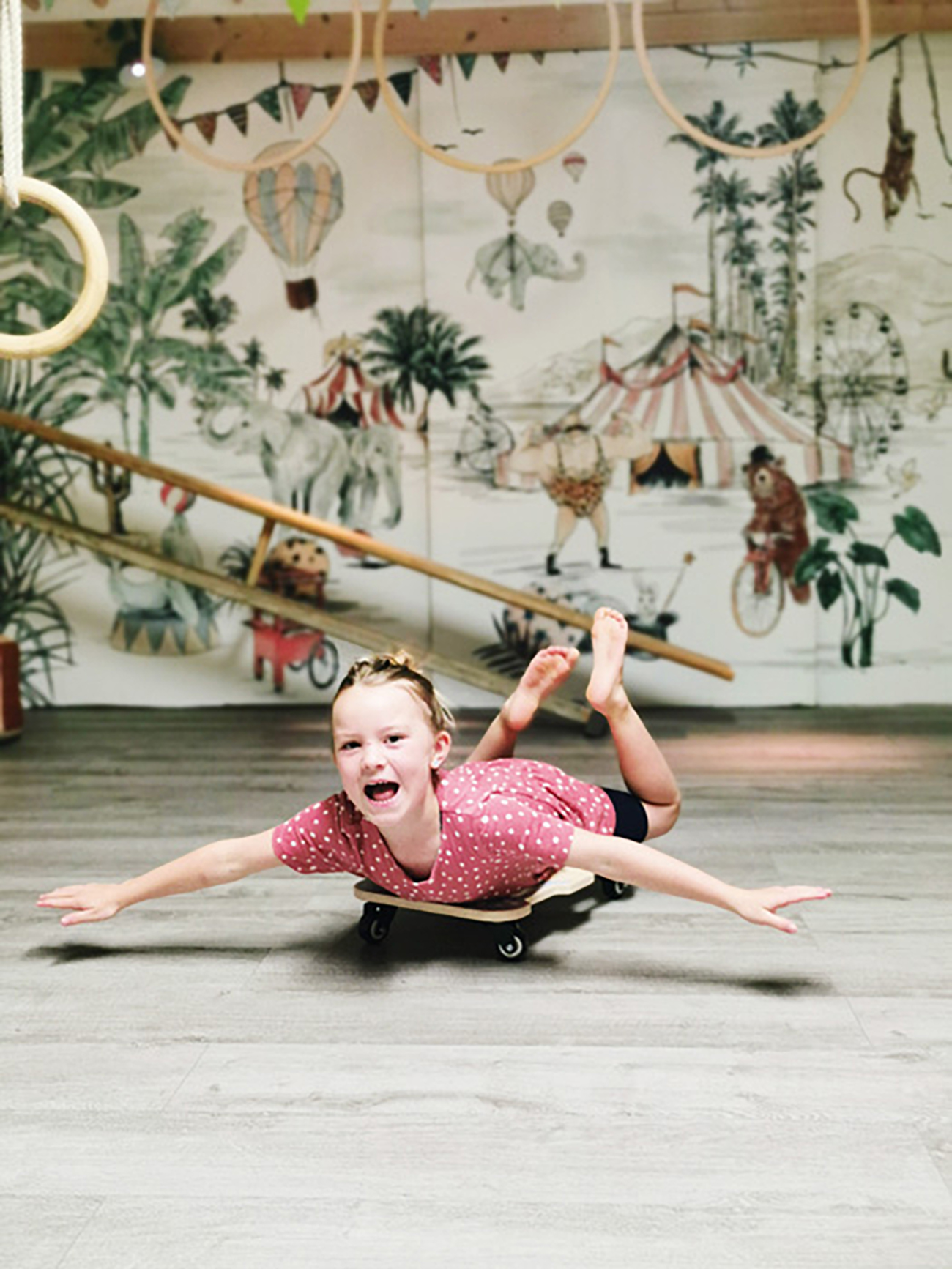 Ein Mädchen auf einem Skateboard in einer Turnhalle, im Hintergrund ein Zirkus-Wandbild.