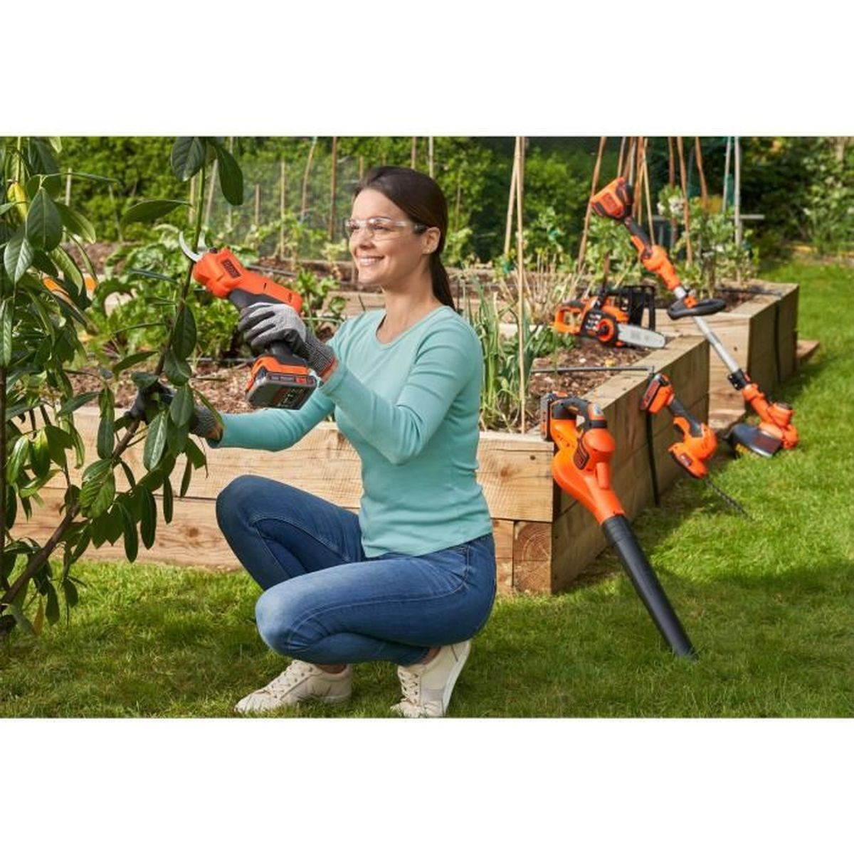 Frau im blauen Hemd hockt mit einer Heckenschere. Andere Werkzeuge und Gartenbeete im Hintergrund.