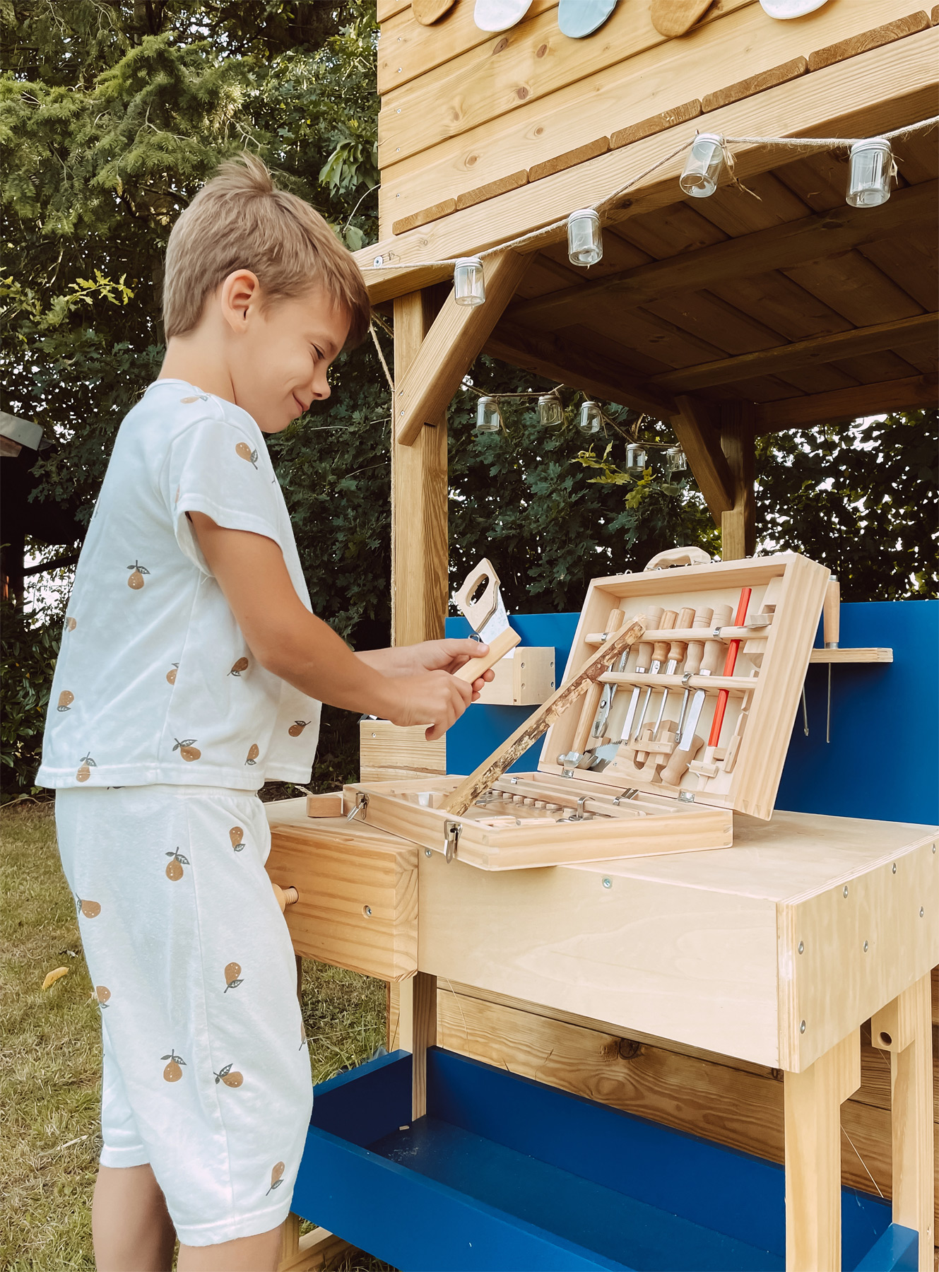 Junge in weißer Kleidung baut mit Werkzeugen. Ein Holzspielhaus steht dahinter.