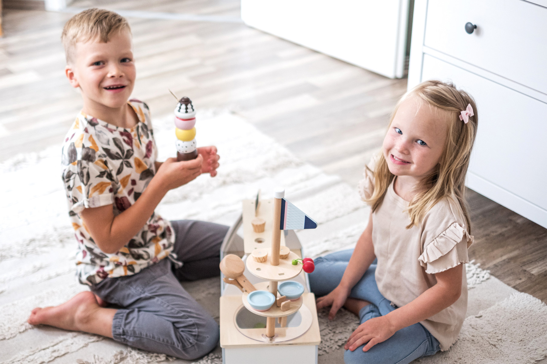 Zwei Kinder lächeln. Ein Mädchen sitzt und ein Junge steht, beide spielen mit Holzspielzeug.