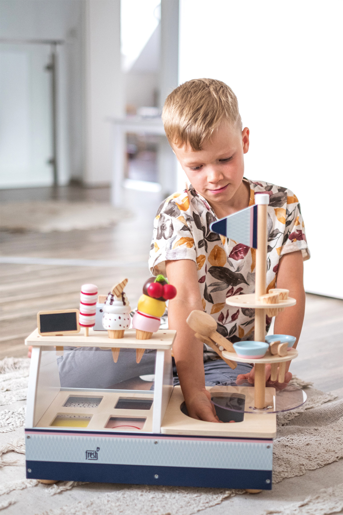 Ein junger Junge spielt mit einem Eisstand aus Holz. Der Stand hat Eistüten und Toppings zur Schau gestellt.