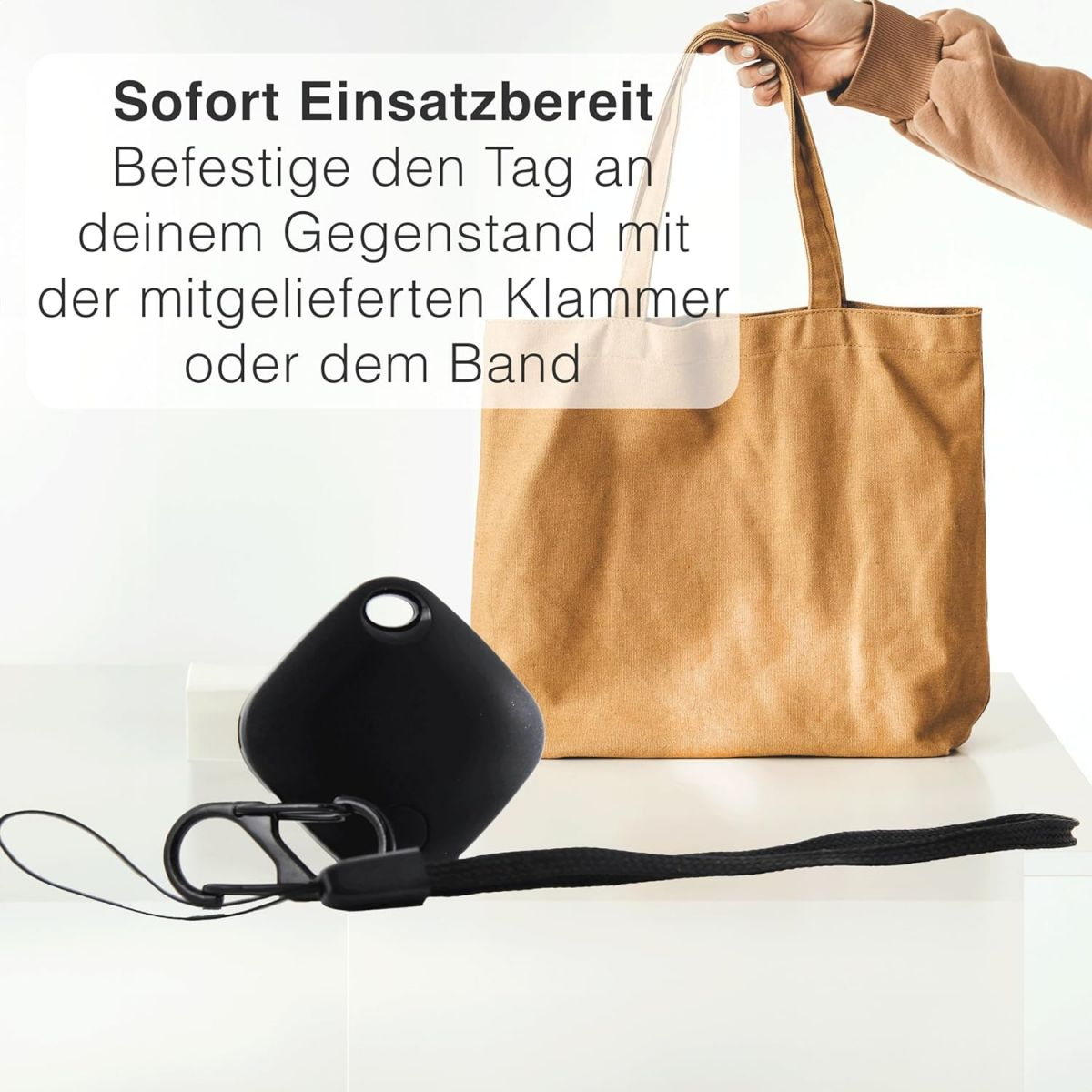 Ein schwarzes Objekt, ein Schlüsselband und eine Tragetasche auf weißer Oberfläche, Hand hält Tasche.
