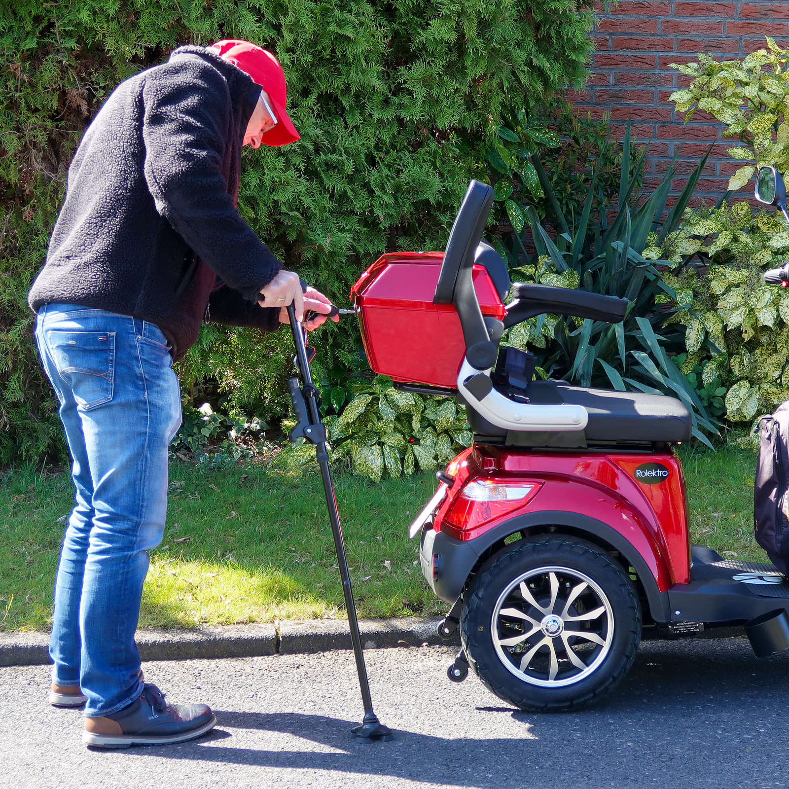 Ein Mann justiert einen Stock neben einem roten Mobilitätsscooter auf einer gepflasterten Straße, grüne Büsche im Hintergrund.