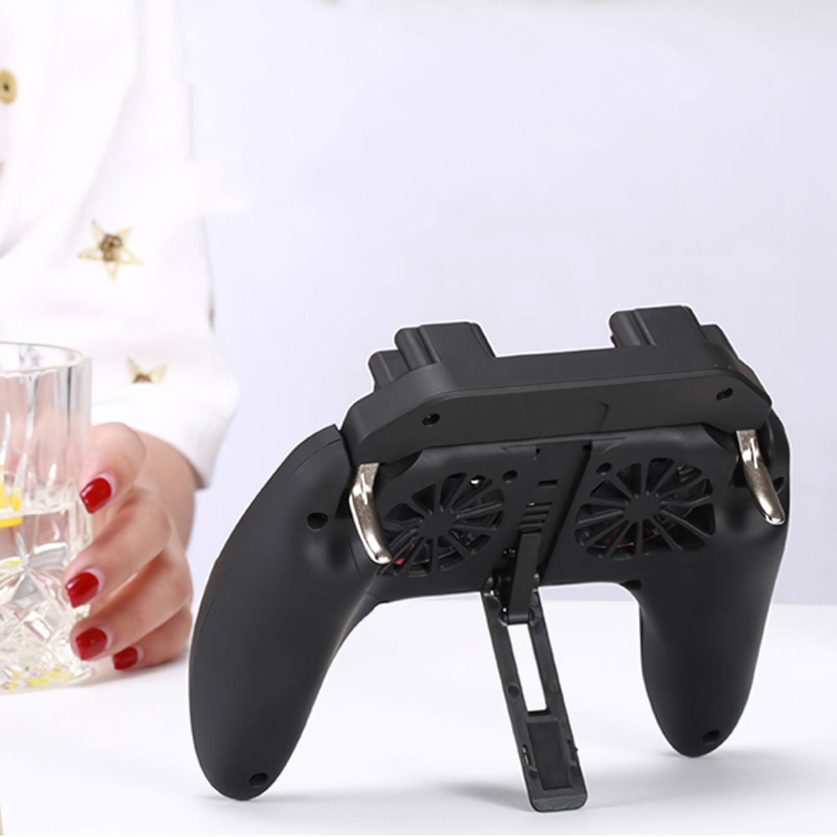 Enfriador de gamepad negro con ventiladores, sostenido por persona con uñas rojas cerca de vaso con agua.