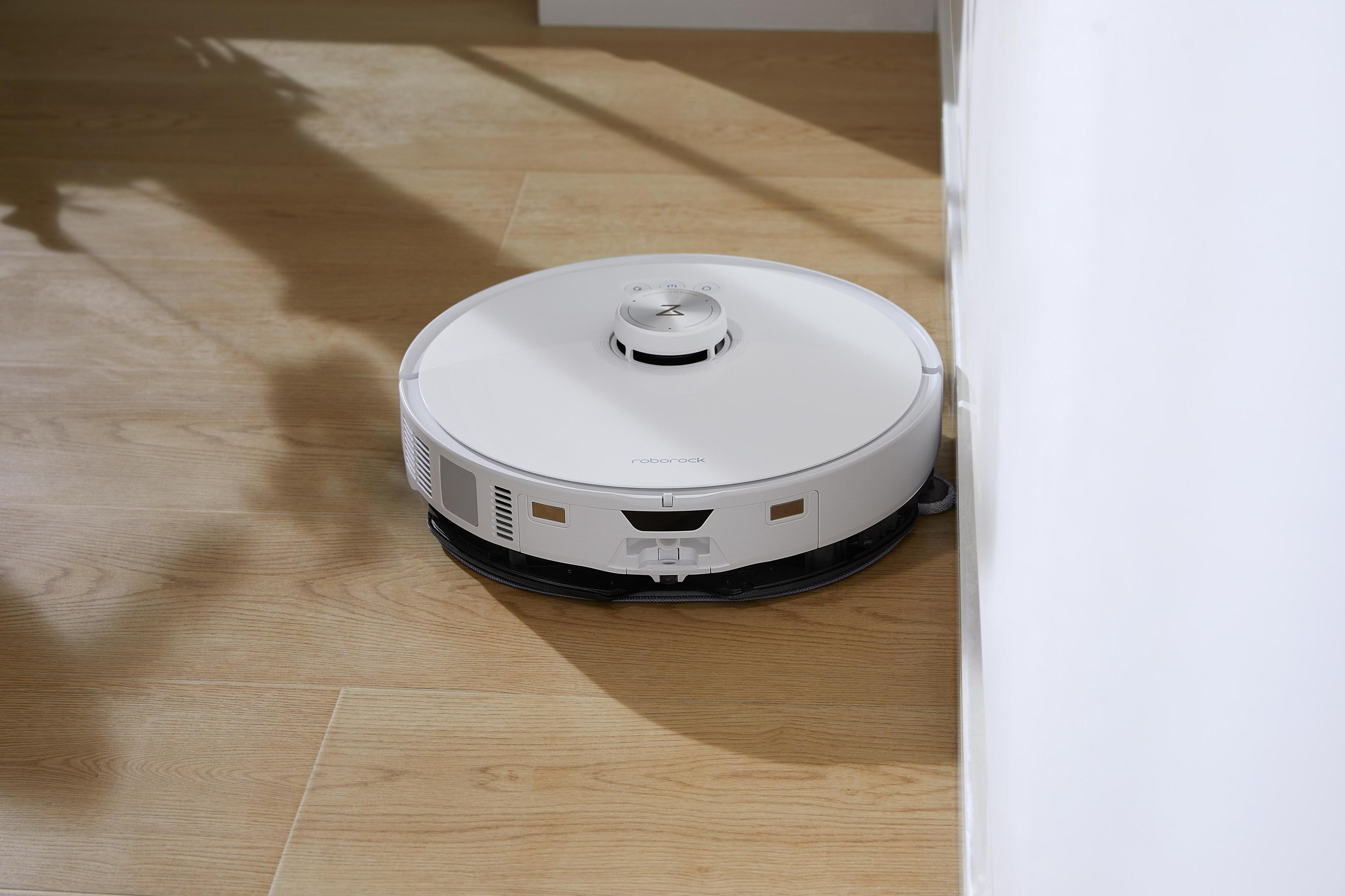 Biały robot sprzątający na drewnianej podłodze. Znajduje się w pobliżu białej ściany i ościeżnicy.