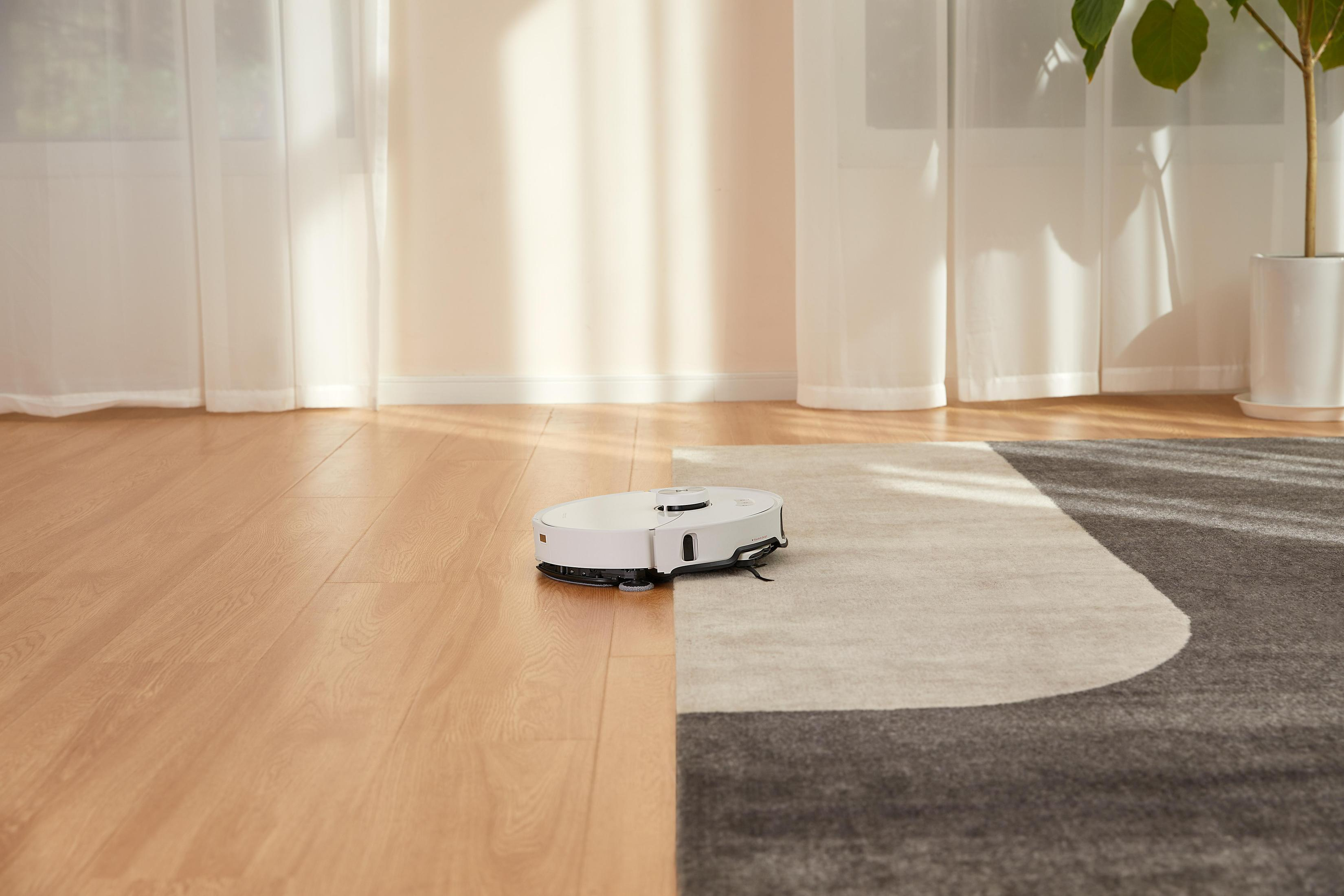 Biały odkurzacz robot na podłodze z drewna i dywanie, blisko okna z zasłonami i rośliną.