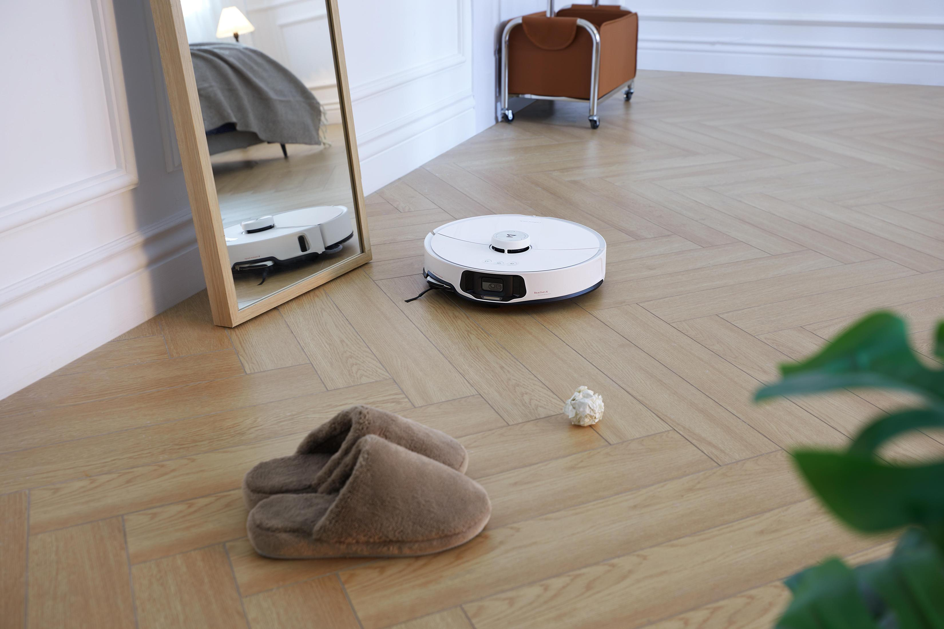 Robot sprzątający na drewnianej podłodze. Lustro odbija go. Kapcie i mała kulka na pierwszym planie.
