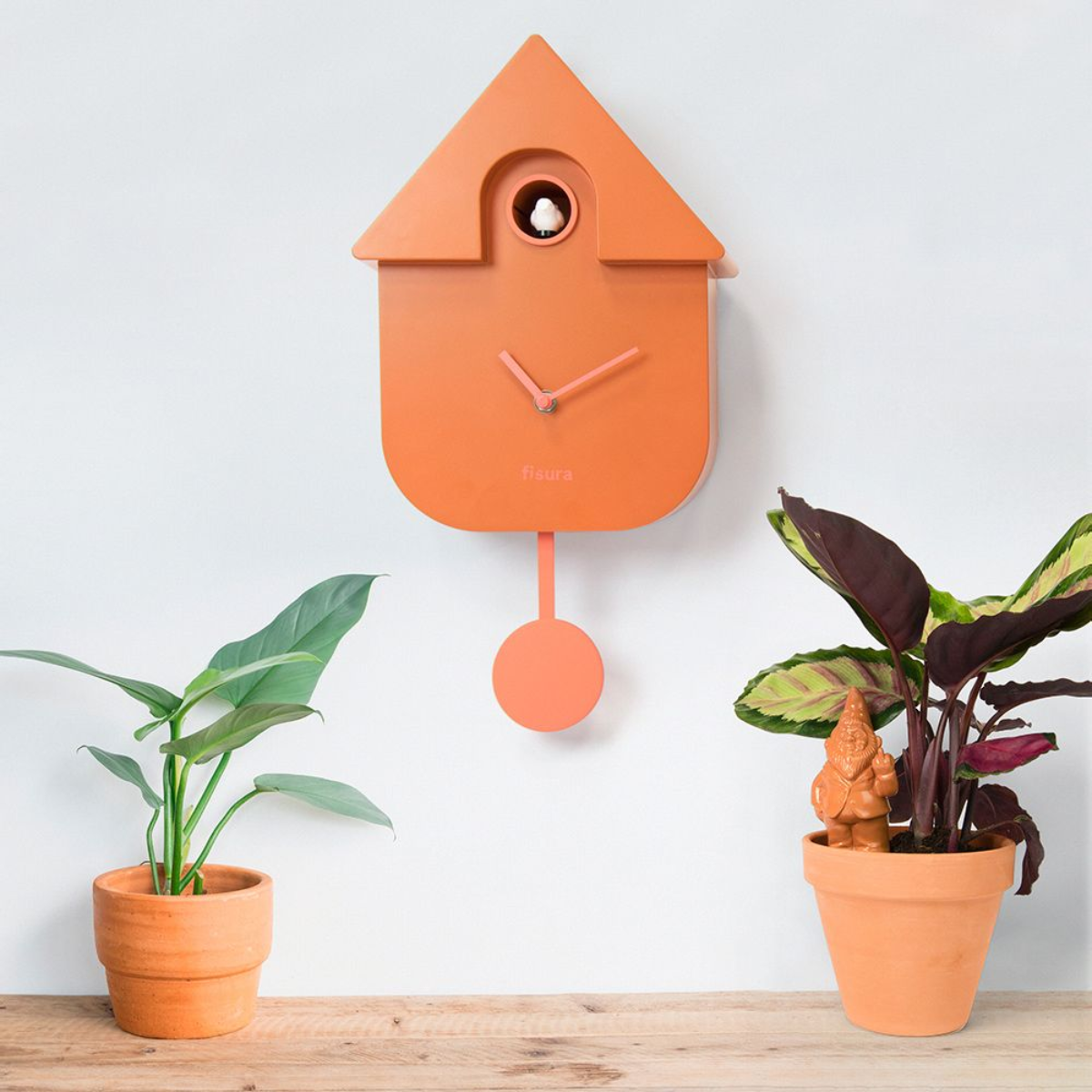 Un orologio a cucù arancione su un muro bianco, con piante in vasi di terracotta.