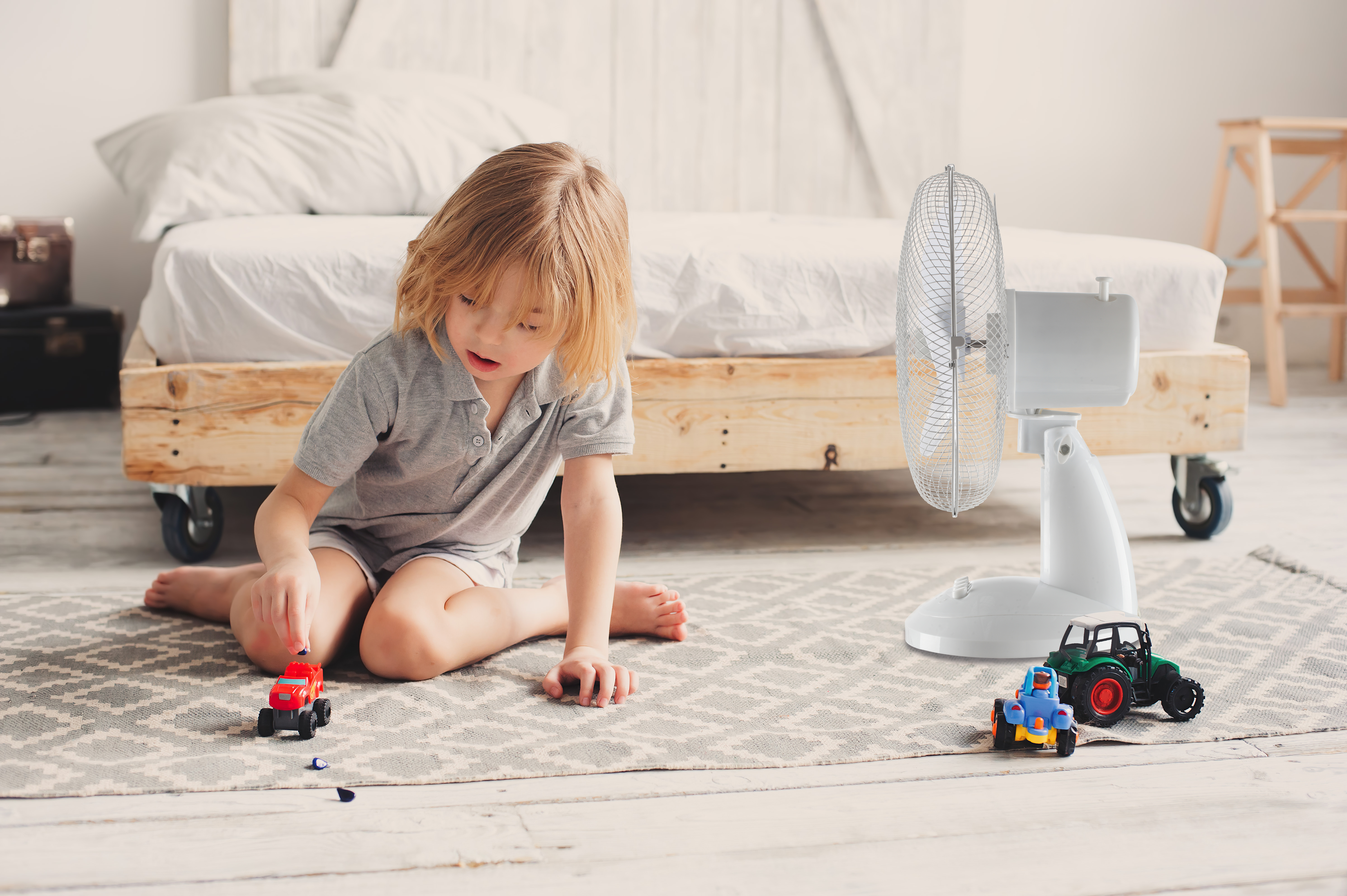Kleinkind spielt mit Spielzeugautos auf einem Teppich. Ein Ventilator und ein Bett sind im Hintergrund des Zimmers.