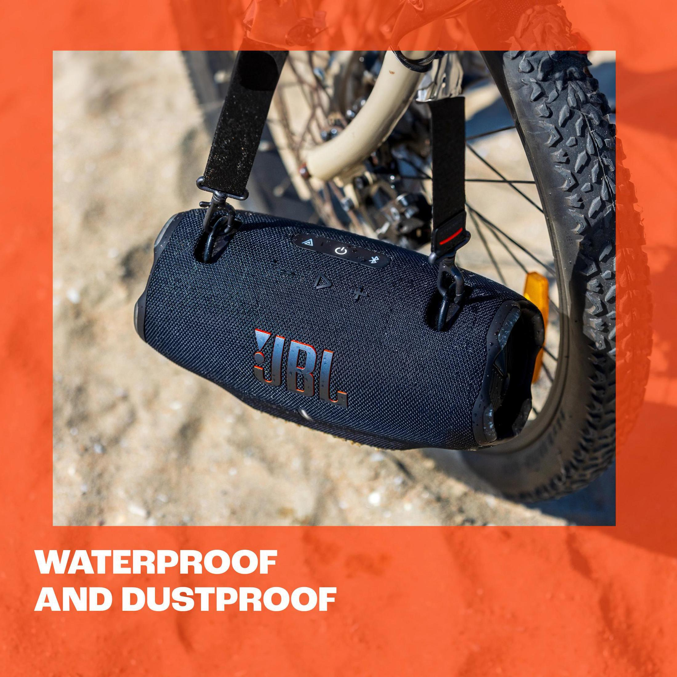 Czarny głośnik JBL z paskiem do noszenia na piaszczystej powierzchni.