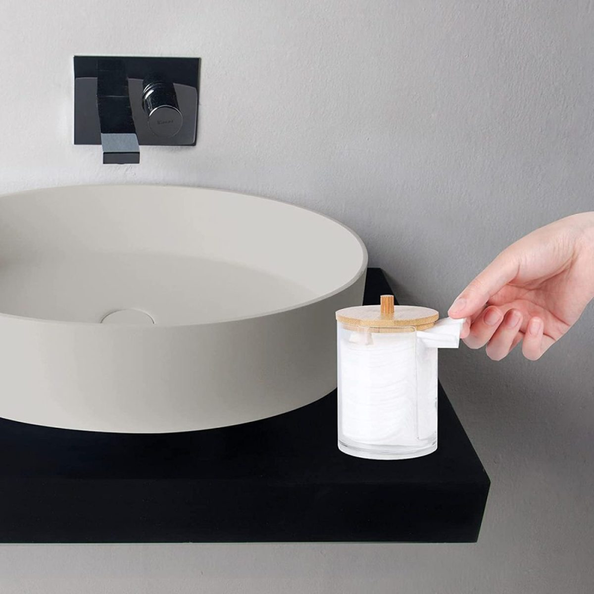 Eine Hand greift nach einem Wattebauschspender. Es ist ein klares Glas mit Holzdeckel, auf einem schwarzen Regal.