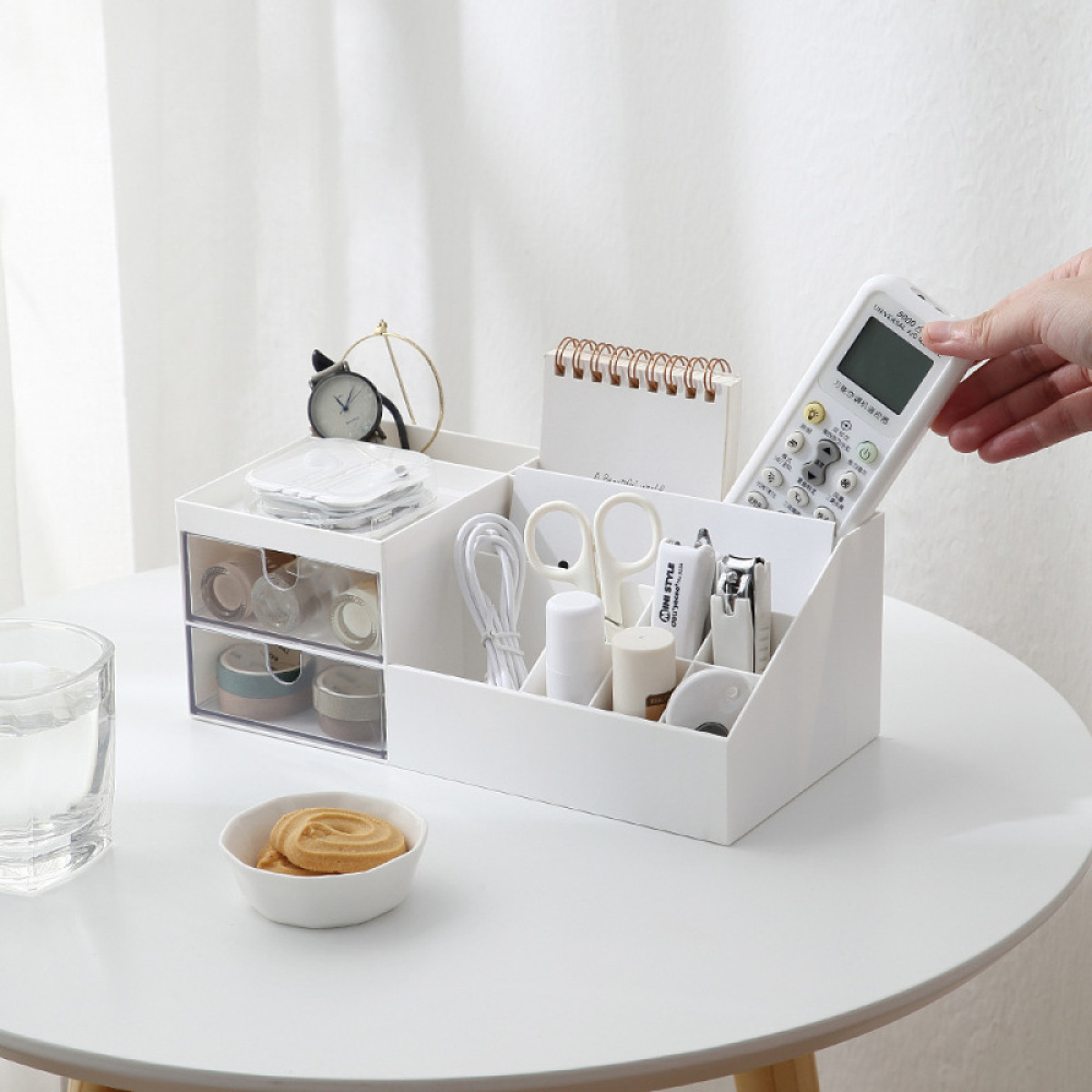 Weißer Schreibtisch-Organizer mit Schere, Fernbedienung und kleiner Uhr auf einem weißen Tisch.