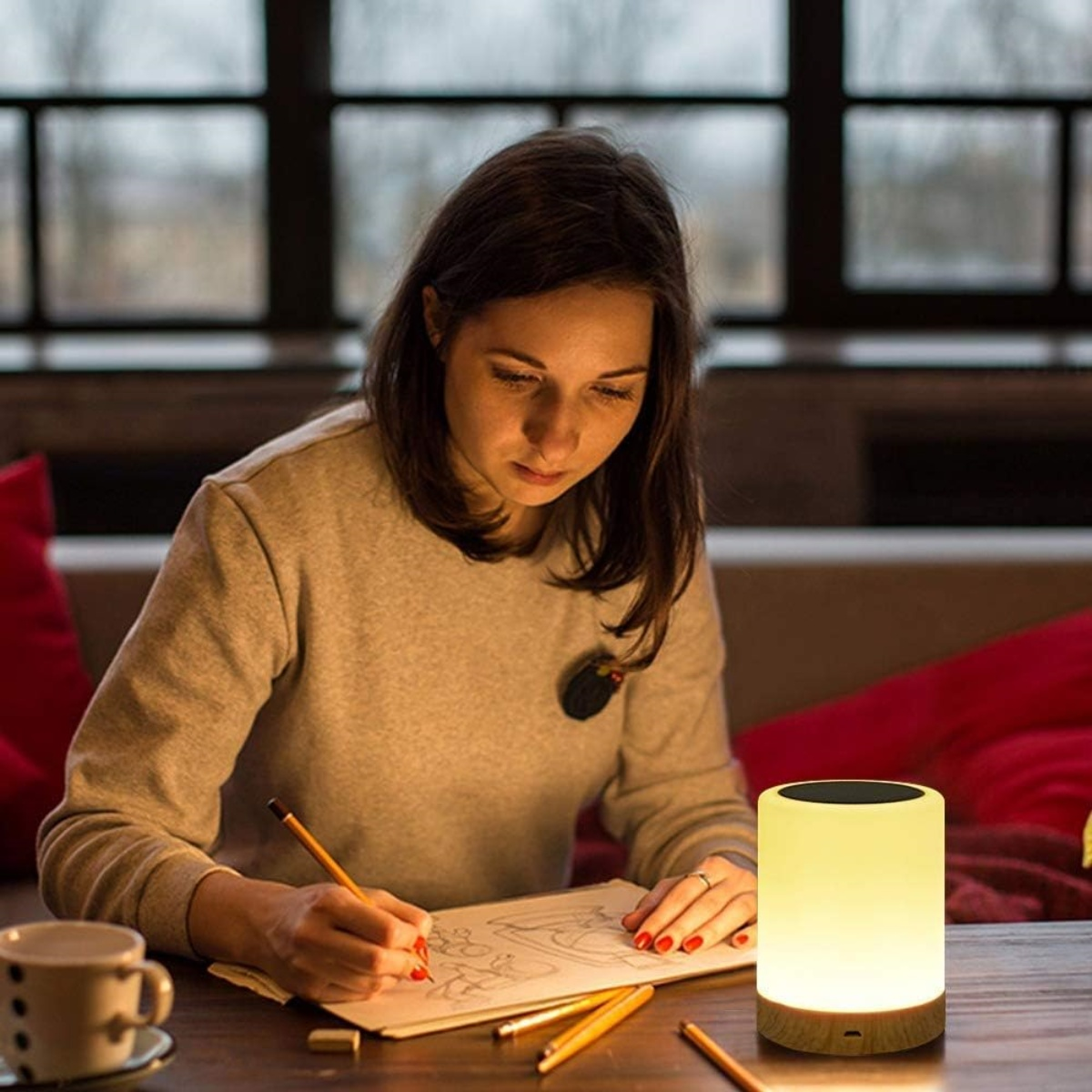 Frau zeichnet an einem Tisch mit einer Lampe. Bleistifte und eine Tasse sind ebenfalls auf dem Tisch.