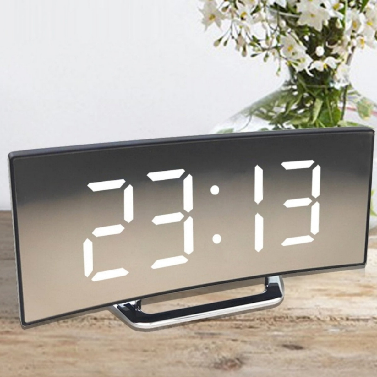 Digitaluhr zeigt 23:13, gewölbte Front, auf Holz. Vase mit weißen Blumen im Hintergrund.